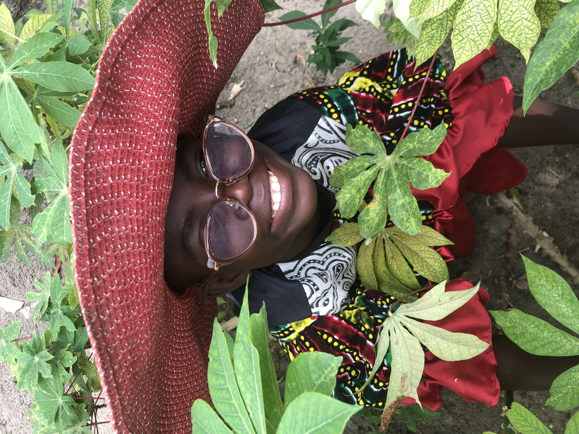 A young person wearing a large red hat and pink-tinted sunglasses smiles widely, surrounded by lush green foliage. They are dressed in a brightly colored, patterned top with red ruffled accents. The background appears to be outdoors, with sandy soil and more green plants visible. The scene is shot from a high angle, looking down at the person.