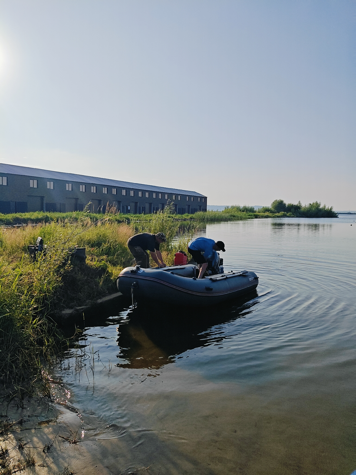 In Chaikovsky, Russia, two men are preparing a gray inflatable boat by the water's edge on a clear, sunny day, likely in the early morning or late afternoon given the low angle of the sun. The boat, which has a red fuel can visible at its rear, is partially submerged in the calm water, with gentle ripples spreading outwards. One man, wearing a dark t-shirt and camouflage pants, is bending over the side of the boat, seemingly making adjustments. The other man, dressed in a blue t-shirt and black shorts, is kneeling inside the boat, facing its rear.
Behind them, a long, modern building with a dark facade and multiple windows stretches horizontally across the landscape. The building is set against a backdrop of lush green reeds and grass, with a line of trees and vegetation visible on the horizon across the water. The sky is a clear, pale blue, with the sun shining brightly in the upper left portion of the image, casting a slight glare. The water is a deep blue-green, reflecting the sky and surrounding environment. The scene appears peaceful and focused on the task of preparing for a boat excursion. There is no visible text in the image.