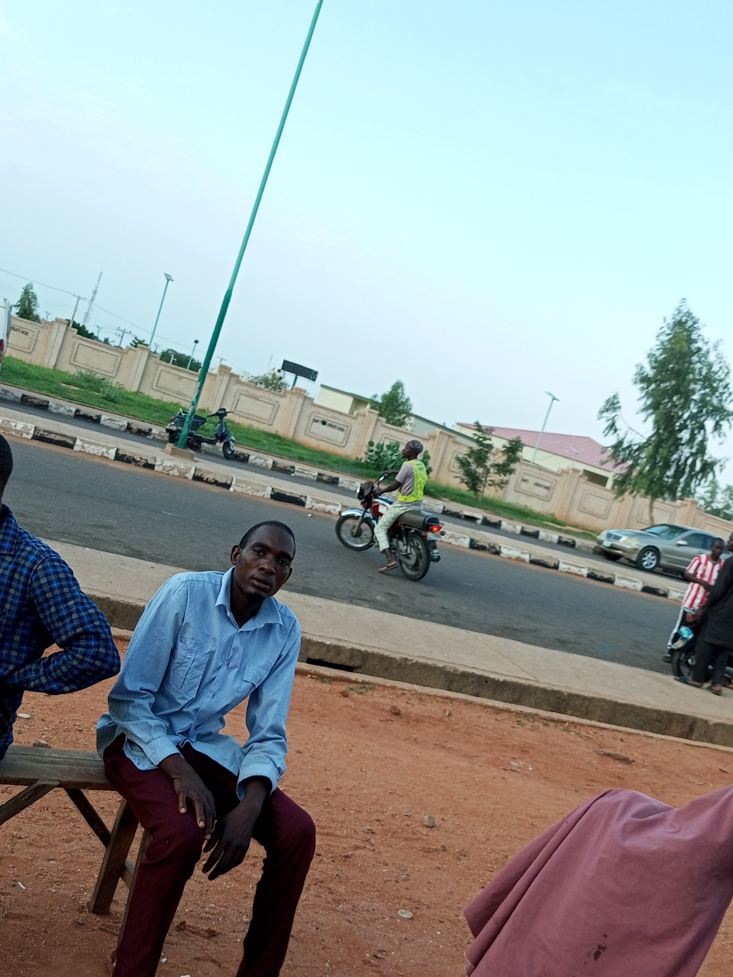 In Gombe, Nigeria, on a street with a concrete curb and asphalt road, two men are visible. One man, wearing a blue collared shirt and maroon pants, sits on a wooden bench in the foreground, looking towards the camera. To his left, another man, seen from the shoulders down, wears a blue and black plaid shirt. Across the street, a man on a motorcycle wears a light green vest and white pants, and a silver car is parked on the side of the road. Further in the background, a dark scooter is parked, and a fence runs along the perimeter of a building. The sky is clear and light blue.