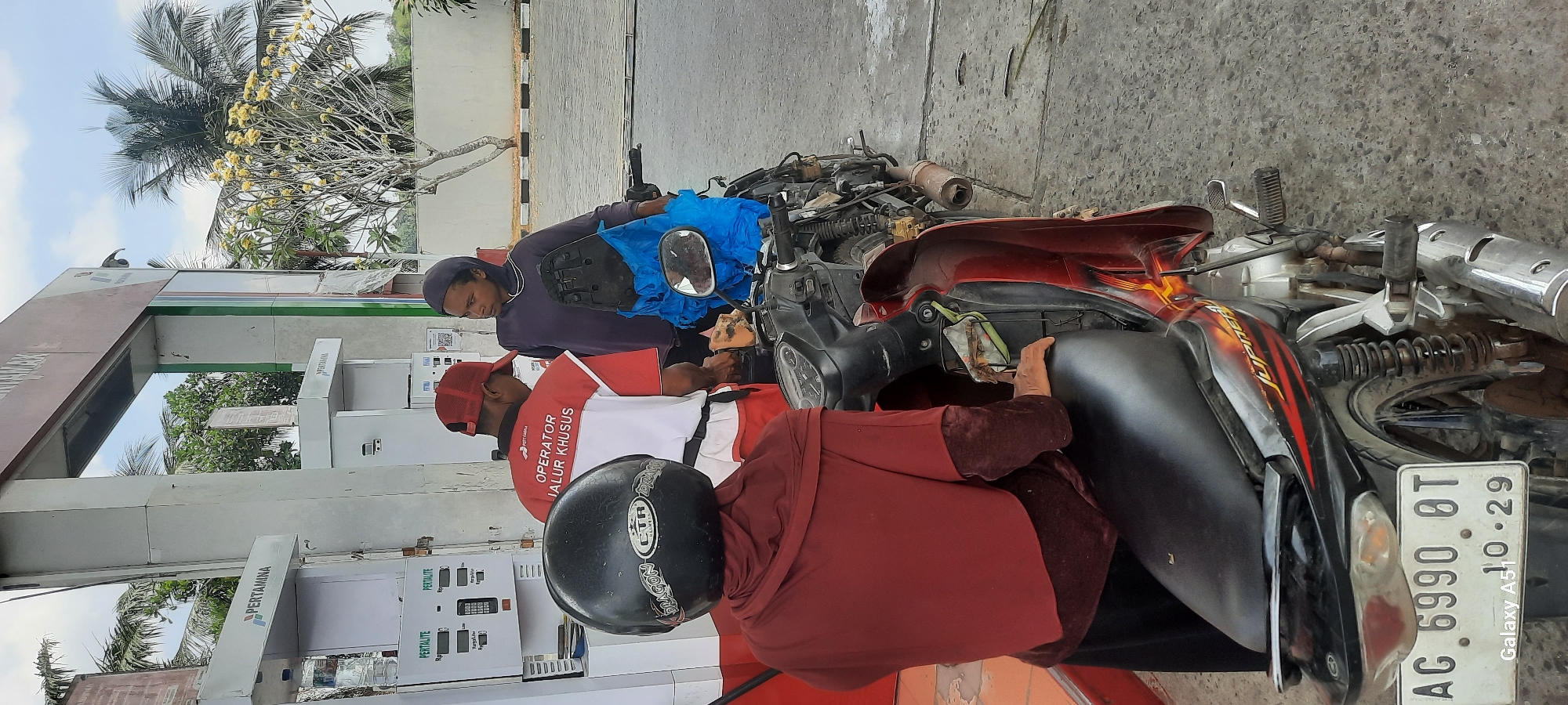 At a Pertamina gas station in Ploso, Indonesia, a person wearing a red hijab and black helmet sits on a red and black motorcycle, facing away from the camera. A gas station attendant in a red and white uniform with "OPERATOR JALUR KHUSUS" (Special Lane Operator) on the back is dispensing fuel into the motorcycle's tank. To the right, another person wearing a dark hoodie and dark hijab looks on. The motorcycle is parked at a gas pump, and the surroundings suggest an outdoor urban environment with palm trees and other vegetation visible in the background. The license plate of the motorcycle is visible and reads "AG 6990 BT".