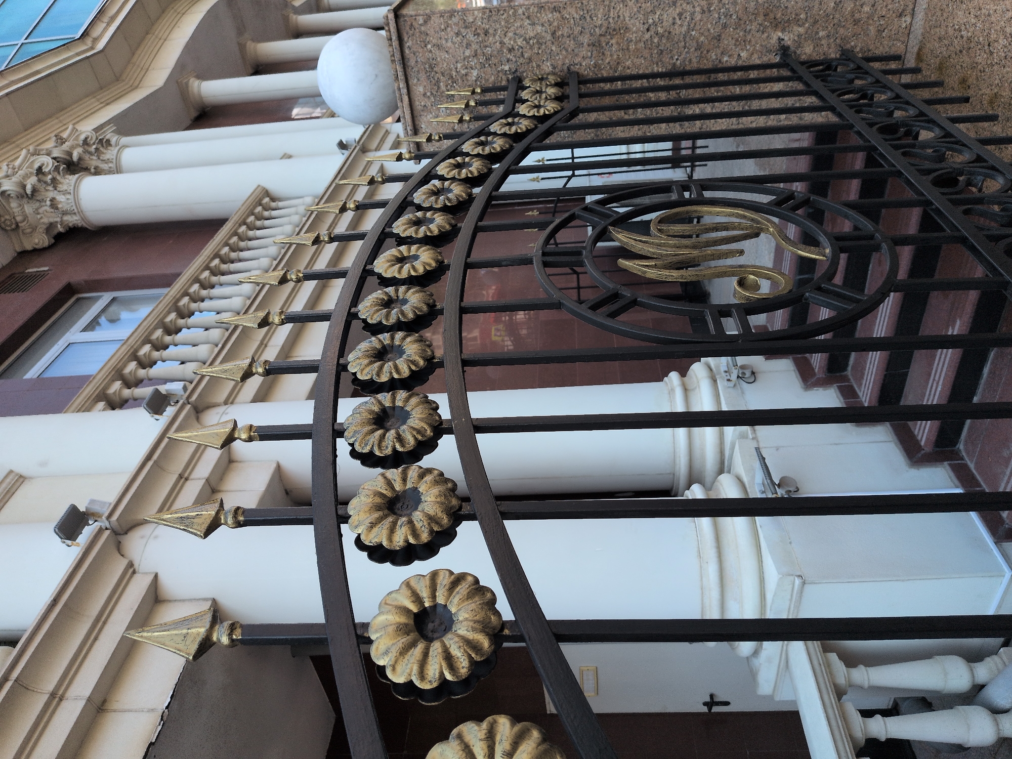 The image shows an ornate black metal gate with golden floral decorations and pointed finials. The gate is in front of a building with white columns, a stone facade, and a balcony. There are two security cameras visible on the building's exterior. In the background, steps lead up to the entrance. The gate features a circular emblem with a golden monogram. The overall scene suggests an upscale or official building.