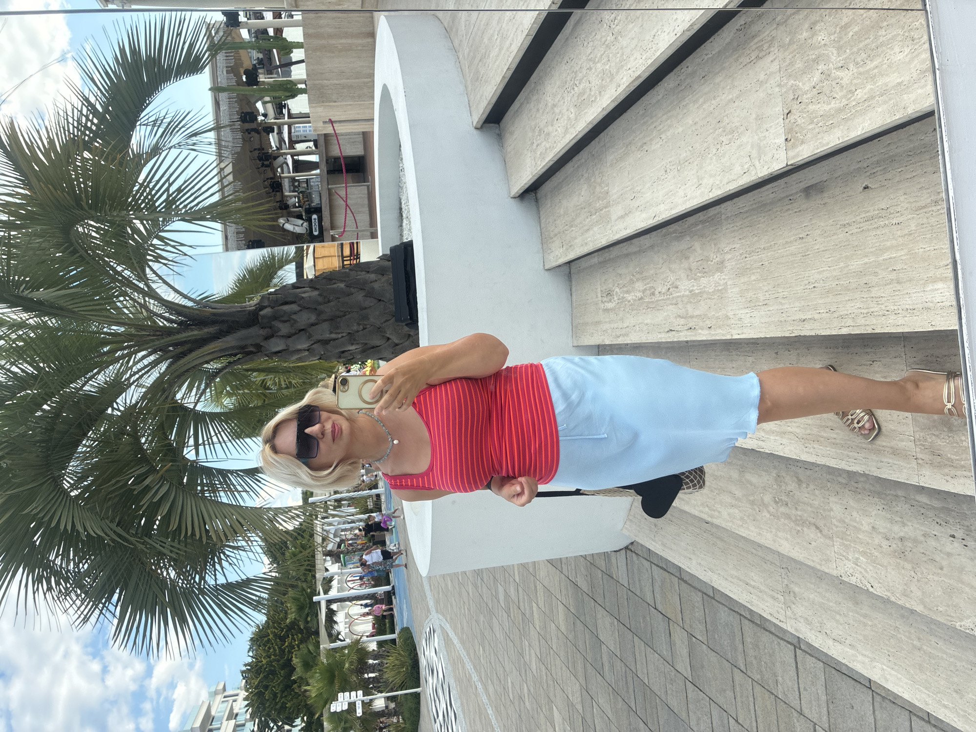 A blonde woman stands on a travertine staircase, taking a selfie in a mirror. She is wearing a pink and red striped tank top, a light blue skirt, gold sandals, and sunglasses. A black bag with a woven pattern hangs from her shoulder. Behind her, a palm tree stands tall, and in the background, there are people walking on a paved area, white structures, and a beach bar with lights. The scene appears to be a modern outdoor venue in Sochi, Russia, possibly a resort or a beach club, given the palm tree and the beach bar. The sky is cloudy.