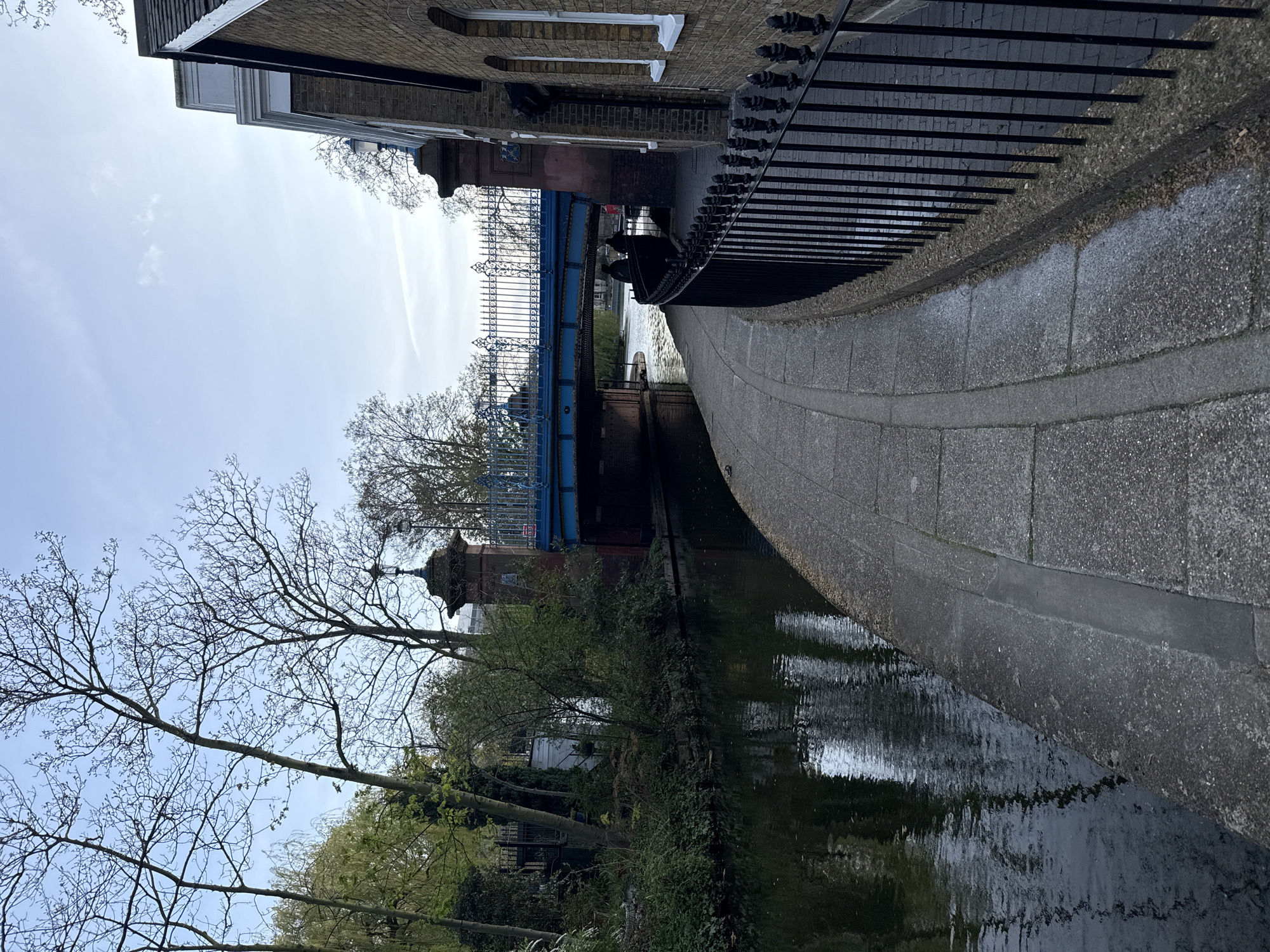 The image shows a canal in London, United Kingdom. A blue bridge with decorative railings spans the canal. A paved path runs alongside the canal, bordered by a black metal fence. Buildings are visible above the fence, including a brick structure with white trim. Trees line the canal bank, some with bare branches. The water of the canal is dark and reflects the surrounding environment. The sky is light blue with some clouds.