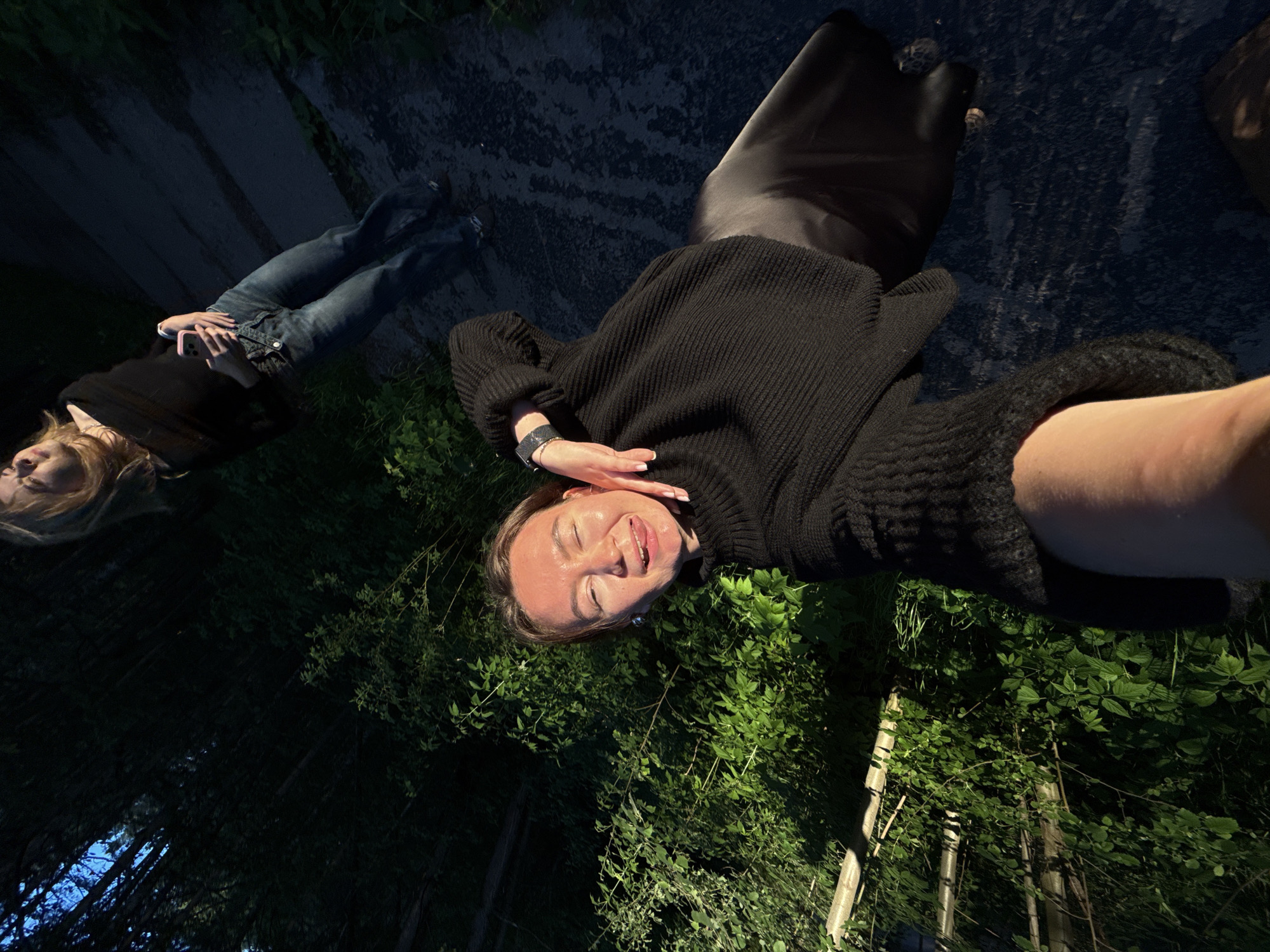 This media file captures two women outdoors in Chaikovsky, Russia, during what appears to be late evening or twilight hours. The image is rotated 90 degrees clockwise, presenting a unique, tilted perspective.
In the foreground, a woman with light brown hair smiles broadly with her eyes closed, suggesting happiness or amusement. She is wearing a black ribbed sweater and a black skirt, with a dark smartwatch visible on her left wrist. Her right hand is gently touching her face, and she appears to be holding the camera to take a selfie.
Behind her, partially out of focus, stands another woman with blonde hair, dressed in a black top and blue jeans. She is holding a smartphone with a pink case in her hands. Both subjects are illuminated by a direct light source, possibly a phone flash, creating highlights on their faces against the darker surroundings.
The setting is outdoors, characterized by abundant dark green foliage, including bushes and trees, which form a dense background. The ground beneath the women appears to be a dark path or pavement. In the deeper background, a glimpse of a dark blue sky and some wooden posts are visible. No discernible text is present in the image. The overall atmosphere suggests a casual, joyful moment between friends in a natural, dimly lit environment.