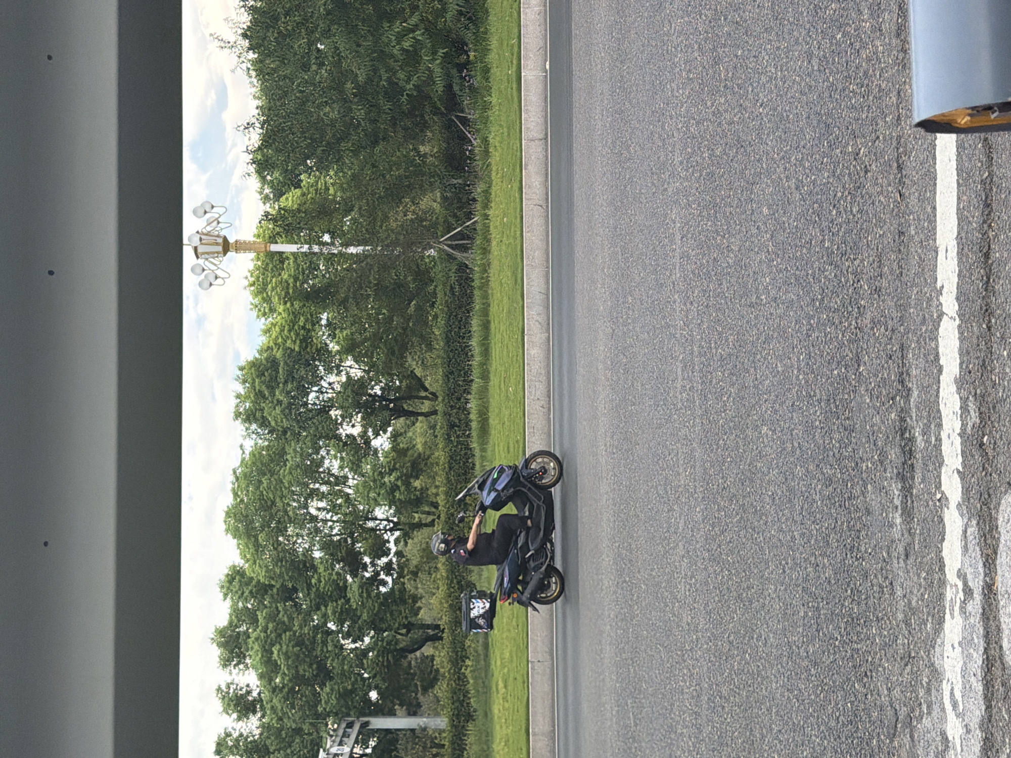 A person wearing a black uniform and a helmet is riding a dark blue scooter on a paved road in Jiangjin, China. The scooter has a black storage box on the back with a white logo. The road is bordered by a grassy median with trees and a lamppost in the background. The sky is cloudy.