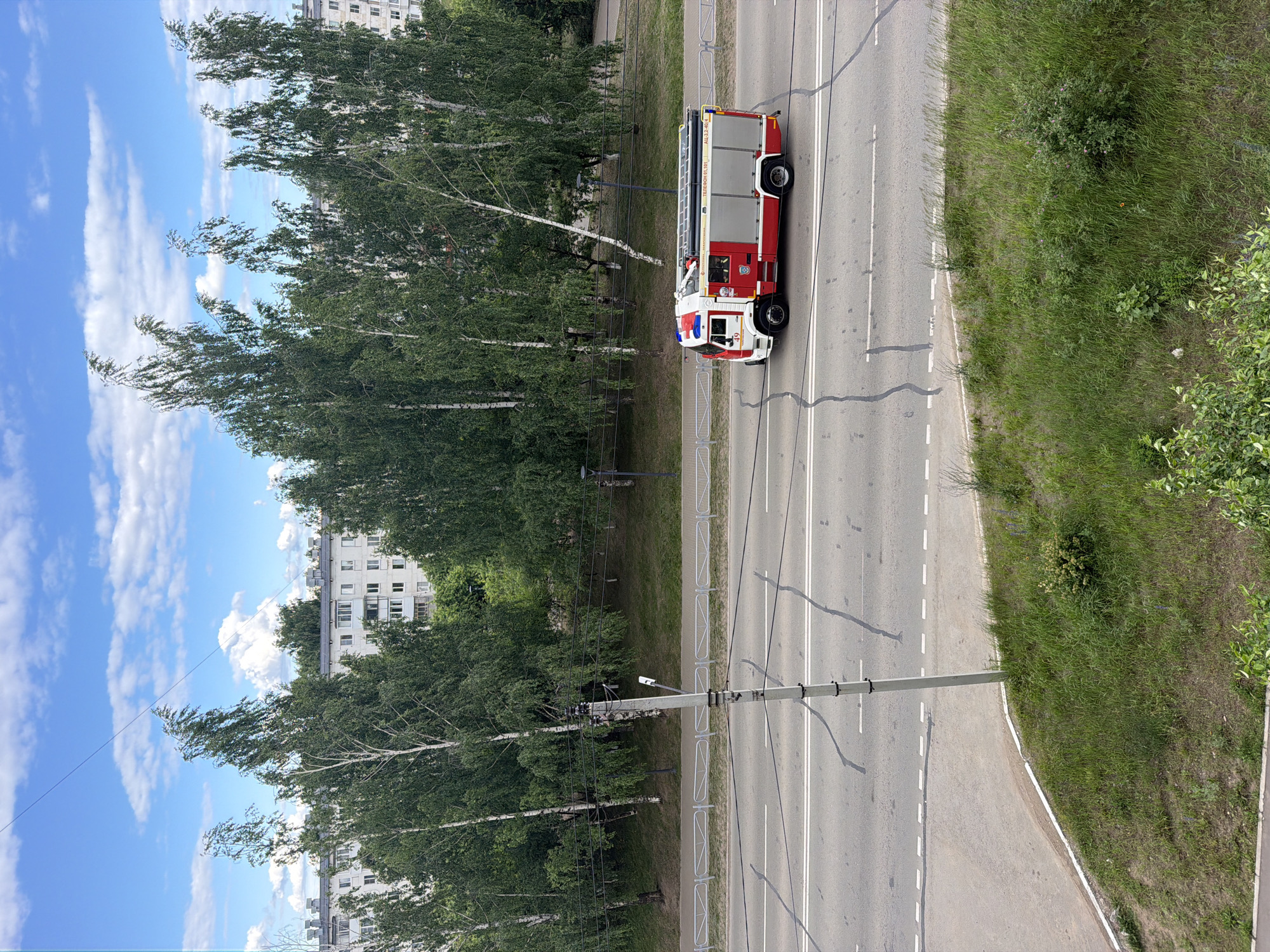 A red and white fire truck with the number 49 is driving on a multi-lane road in Chaikovsky, Russia. The fire truck has a ladder rack on its roof and markings on its side. The road has white lane dividers and a light pole with wires is situated on the side of the road. Behind the road, there is a grassy embankment with trees and residential buildings in the background. The sky is blue with scattered white clouds.