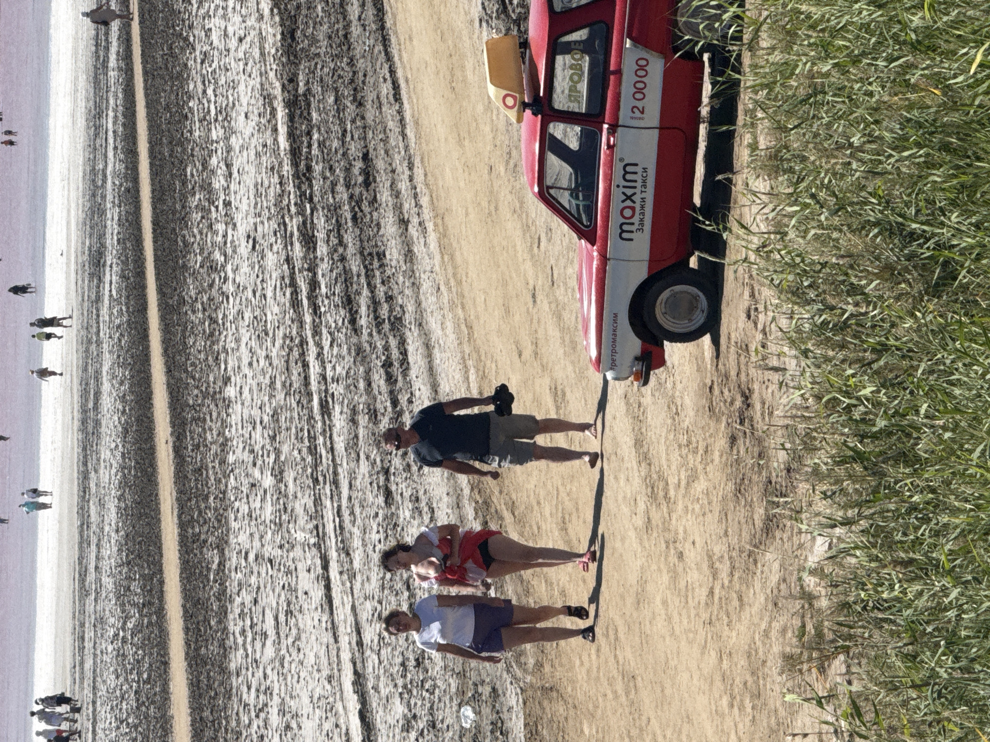 The image depicts a scene at the mud baths of the salt lake in Bursol, Russia. In the foreground, three people are walking on a sandy path. From left to right, a woman in a white t-shirt and purple shorts is walking, followed by a woman in a red and white outfit, and then a man in a dark t-shirt and grey shorts. To the right, a red vintage taxi with a yellow taxi sign on its roof is parked. The taxi has the text "maxim Закажи такси" on its side, along with a phone number. The background shows a vast expanse of muddy terrain, with several people scattered in the distance, appearing to be at the mud baths. Tall, green reeds are visible in the bottom right corner of the image. The sky is a pale pinkish-purple, suggesting it might be sunset or sunrise.