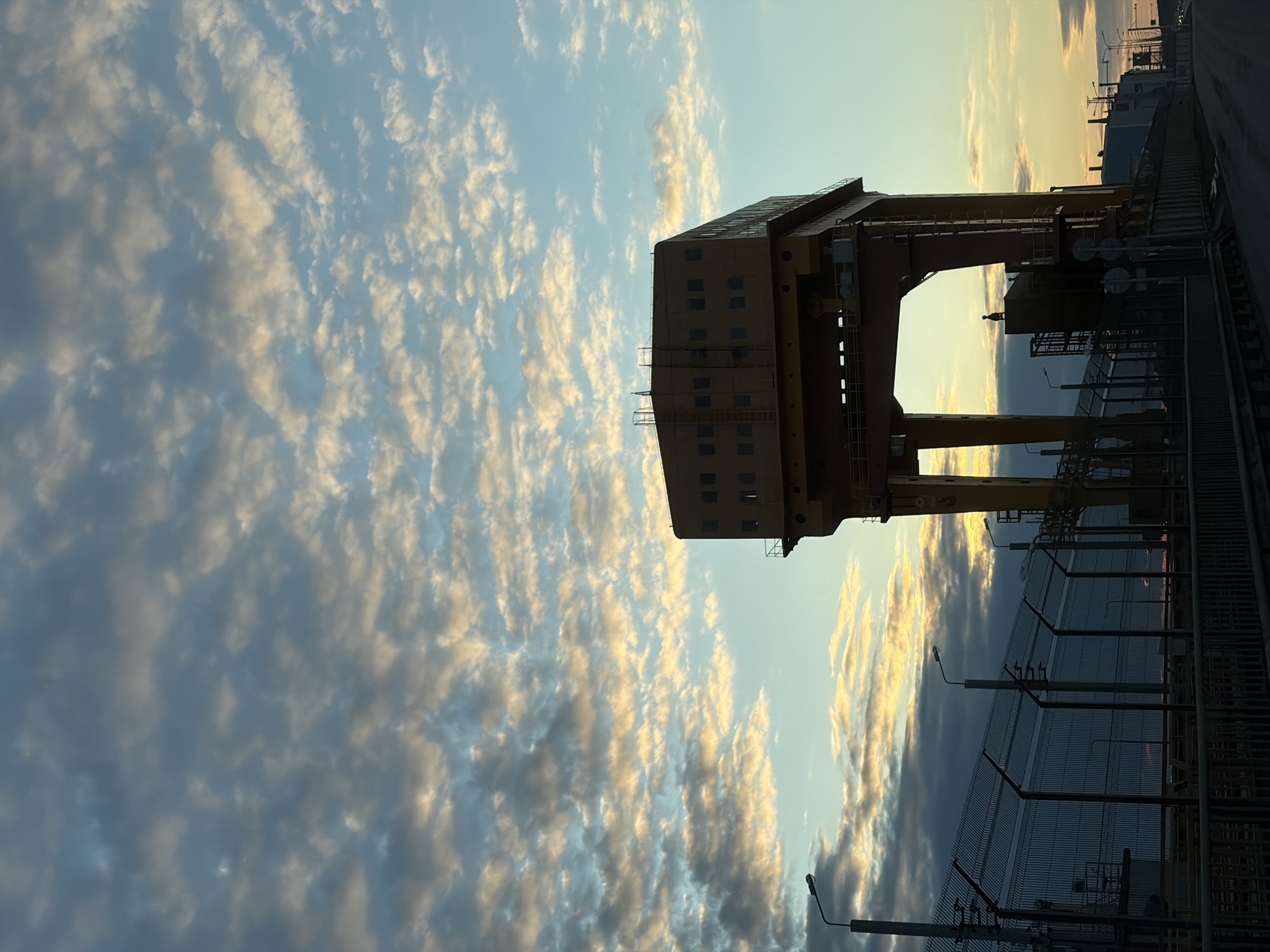 The image shows a large, industrial-looking structure in Chaikovsky, Russia. The structure is tall and yellow, with a rectangular building-like section at the top, supported by two thick columns. There is metal fence and other industrial equipment near the structure. The sky is filled with clouds that are lit by the rising or setting sun. The overall scene is somewhat surreal due to the angle of the photo.