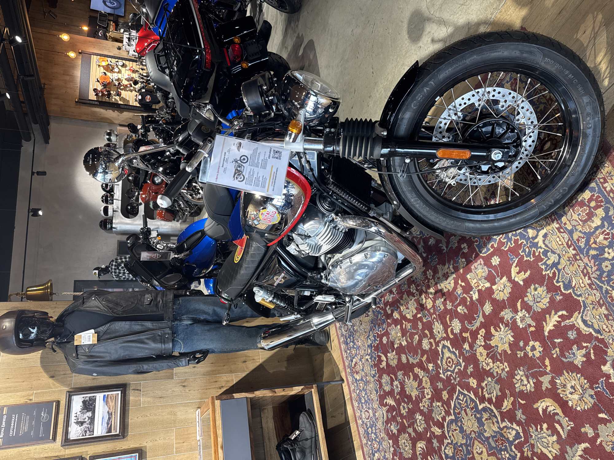 Here's a detailed description of the image:
The image shows a display inside what appears to be a motorcycle dealership, potentially in Saint Petersburg, Russia, based on the Cyrillic text present on signage.
**Subjects and Objects:**
* **Motorcycles:** Several motorcycles are visible. The primary focus is a black Royal Enfield Interceptor 650. Other motorcycles, including blue and black models, are partially visible in the background.
* **Motorcycle Tire:** The front tire of the Interceptor is prominently displayed. The tire brand is CEAT, size 100/90-18 M/C 56H.
* **Mannequin:** A mannequin dressed in a black leather jacket, blue jeans, and a helmet is positioned next to a wooden display case.
* **Sales Information:** A display card is attached to the Royal Enfield with the motorcycle's specifications and price (699,000P, likely rubles).
* **Documents:**Framed documents are on the wall, including a Royal Enfield certificate.
* **Wooden Display Case:** A wooden display case features motorcycle boots.
**Environment:**
* **Flooring:** The foreground features a red rug with floral patterns. The rest of the flooring is wood laminate.
* **Walls:** Walls are wood paneled, adding to the dealership's aesthetic.
* **Lighting:** There are lights present on the ceiling.
**Additional details**:
* The scene appears to be a showroom or retail space displaying and advertising motorcycles and associated gear.
* The brand Royal Enfield is heavily featured.