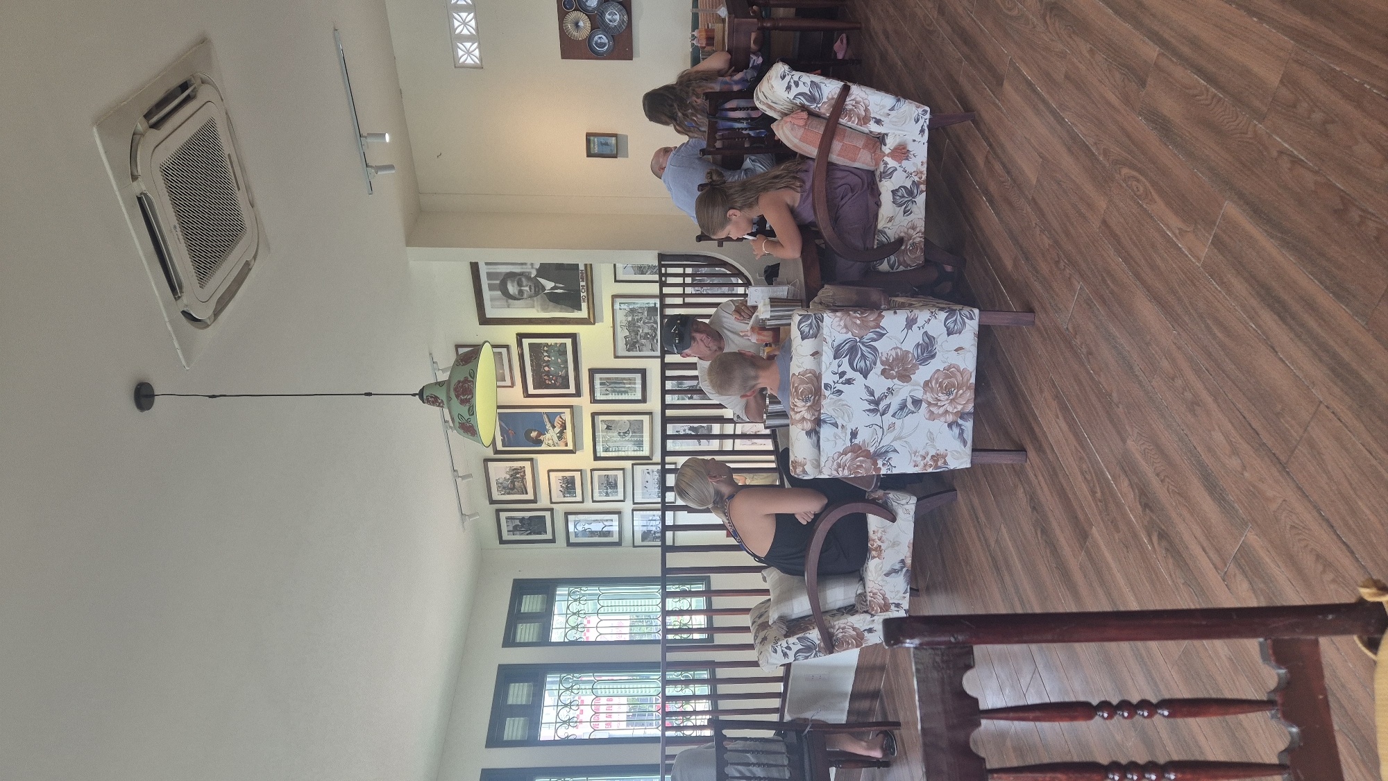The image depicts the interior of a brightly lit cafe, likely in the city of Đà Nẵng, Vietnam, as evidenced by the portrait of Ho Chi Minh and the overall aesthetic which is typical of Vietnamese cafes. Several groups of people are seated at tables, engaged in conversation and eating. In the foreground, a woman with blonde hair in a black dress is seated in a floral armchair, facing away from the camera and talking to a young boy. Next to them, a man wearing a black cap and a white t-shirt is also seated at a table. To the right, another young girl in a purple dress is seated in a similar floral armchair. In the background, other patrons are visible at tables, including a man in a blue shirt and a woman with long brown hair. The cafe walls are adorned with numerous framed black and white photographs and portraits, including a prominent portrait of Ho Chi Minh. A pendant lamp with a floral shade hangs from the ceiling, and an air conditioning unit is installed above. The flooring is made of wooden planks.