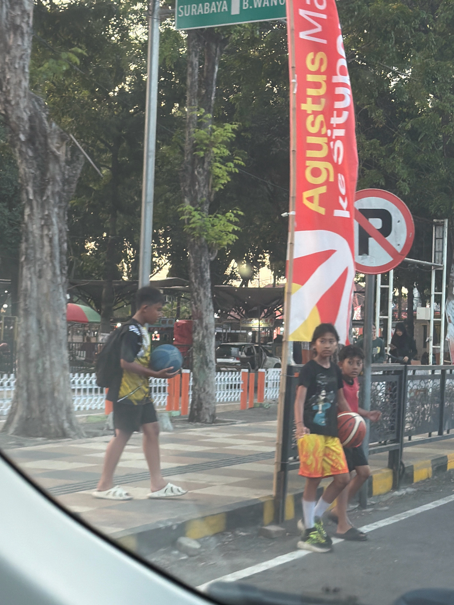 In Situbondo, Indonesia, on a paved sidewalk next to a street, two young boys are walking. The boy on the left, wearing a black and yellow shirt and black shorts, carries a black backpack and a blue basketball. The boy on the right, wearing a black t-shirt and yellow and orange patterned shorts, carries an orange basketball. A red and white banner with Indonesian text, including "Agustus" and "Situbondo," is visible. A "No Parking" sign is also present. Trees line the sidewalk, and a white fence separates the sidewalk from other areas. The scene appears to be during daylight hours.