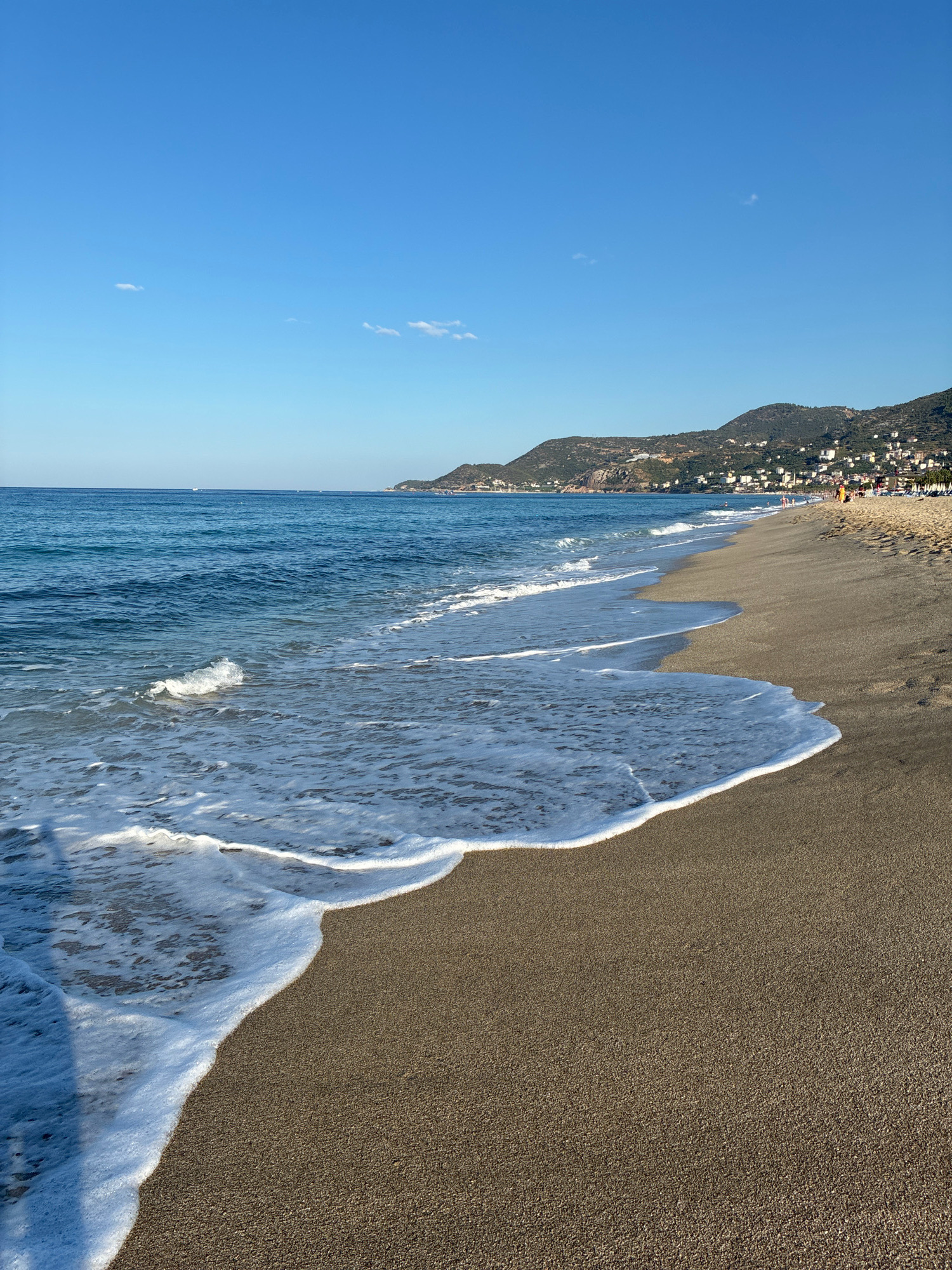 The image captures a sunlit scene at Cleopatra Beach in Alanya, Turkey. The foreground is dominated by the wet, dark sand of the beach, where a gentle wave is receding, leaving behind a foamy white line of sea spray. The water is a deep blue, with smaller waves and ripples extending to the horizon. In the distance, a coastline rises, covered in lush green vegetation and dotted with white buildings that climb the hillsides. A clear, bright blue sky stretches above, with a few wispy white clouds. Several people are visible in the distance on the beach, some walking and others near beach chairs or umbrellas. The overall impression is one of a serene and inviting beach environment on a beautiful day.