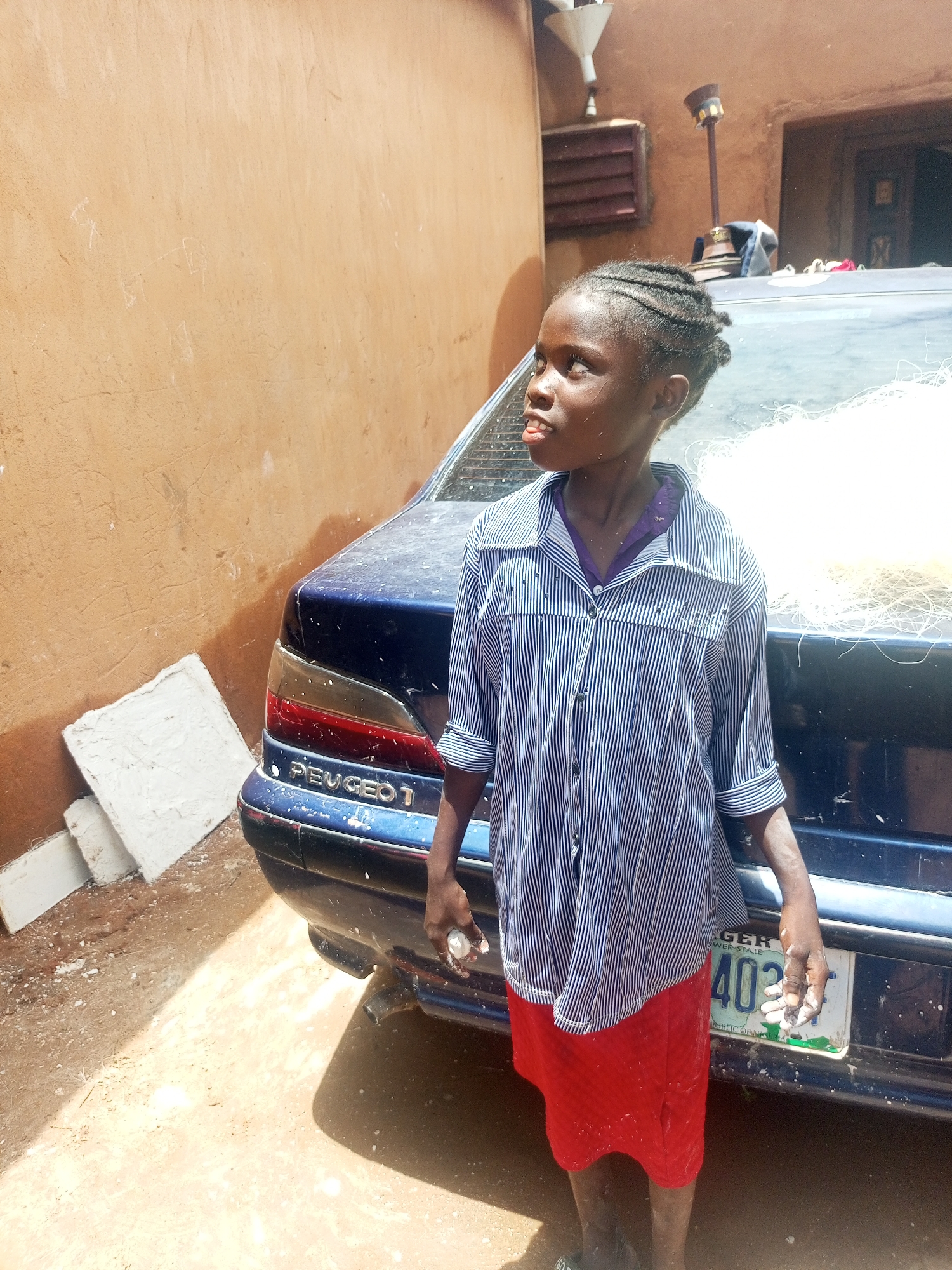 In Gombe, Nigeria, a young person stands next to a blue Peugeot sedan in an outdoor, sandy environment. The person is wearing a striped button-down shirt and a red skirt. Their hair is styled in braids. The car's license plate is visible, and there is some white material on the car and on the person's hands. The background shows a textured, orange-brown wall and a doorway. Several white rectangular objects are stacked near the car.