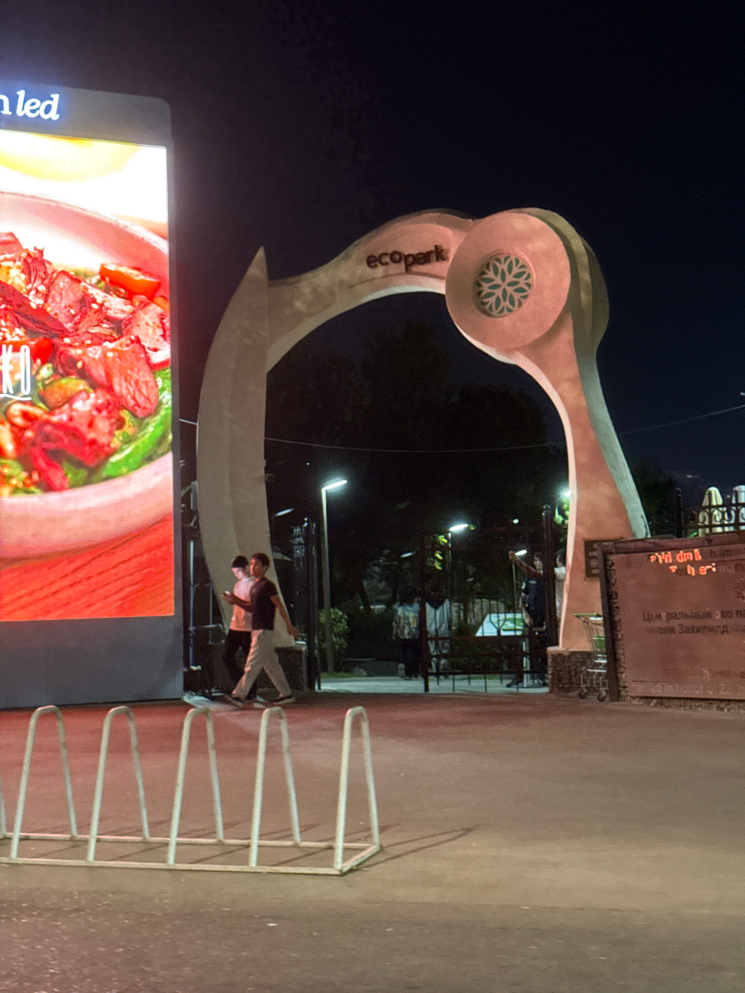 The image captures a nighttime scene at the entrance of Ecopark in Tashkent, Uzbekistan. To the left, a large LED screen displays an advertisement featuring a close-up of a dish with meat, vegetables, and sesame seeds. The words "n led" are visible at the top of the screen.
Dominating the center of the frame is a large, stylized archway that forms the entrance to the park. The arch is made of a light-colored material and features a circular decorative element with a green floral pattern. The word "ecopark" is prominently displayed on the arch.
In front of the archway, two young men are walking towards the park entrance. The man on the left is wearing a white t-shirt and dark pants, while the man on the right is in a dark t-shirt and light-colored pants. Both are looking at their phones.
A metal bicycle rack with several slots is positioned in the foreground, facing the entrance. The ground is paved, and a streetlamp with a modern design is visible to the right of the archway, illuminating the area.
To the right of the arch, a large, weathered sign with text in red and white letters is partially visible. The red text appears to be in a foreign script. Below this, there is a darker sign with Cyrillic lettering, suggesting the presence of information or a memorial. A shopping cart is visible near the right edge of the frame. The overall environment suggests an urban public space at night.