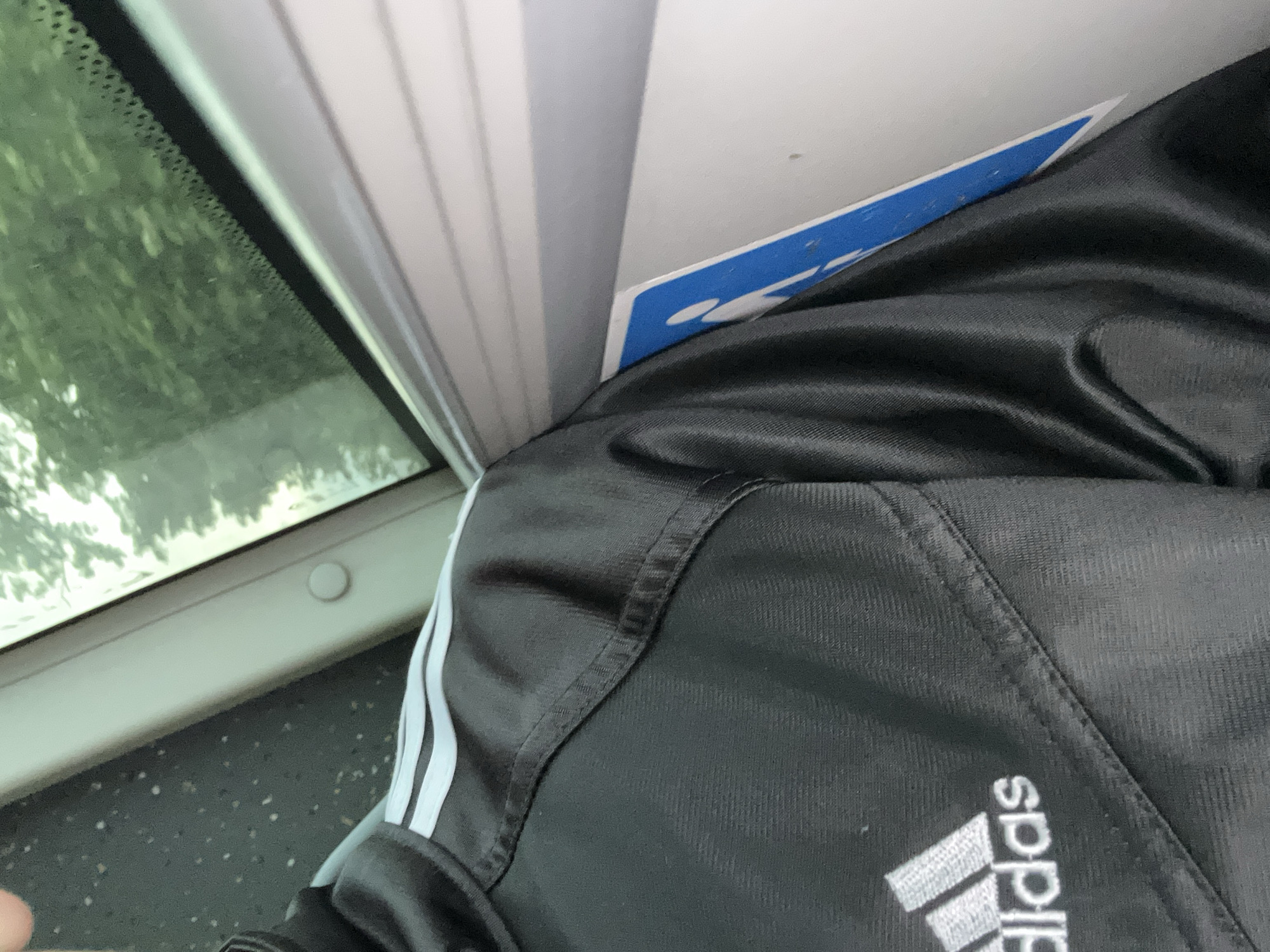 The image shows a close-up view of a person wearing a black Adidas jacket with white stripes on the shoulder. The jacket has a shiny texture and is stitched in diagonal patterns. A blue and white sticker with a pictogram of a person walking is partially visible on the right side of the image. In the background, a window reveals blurred green foliage, suggesting an outdoor environment. The lighting is natural and diffused.