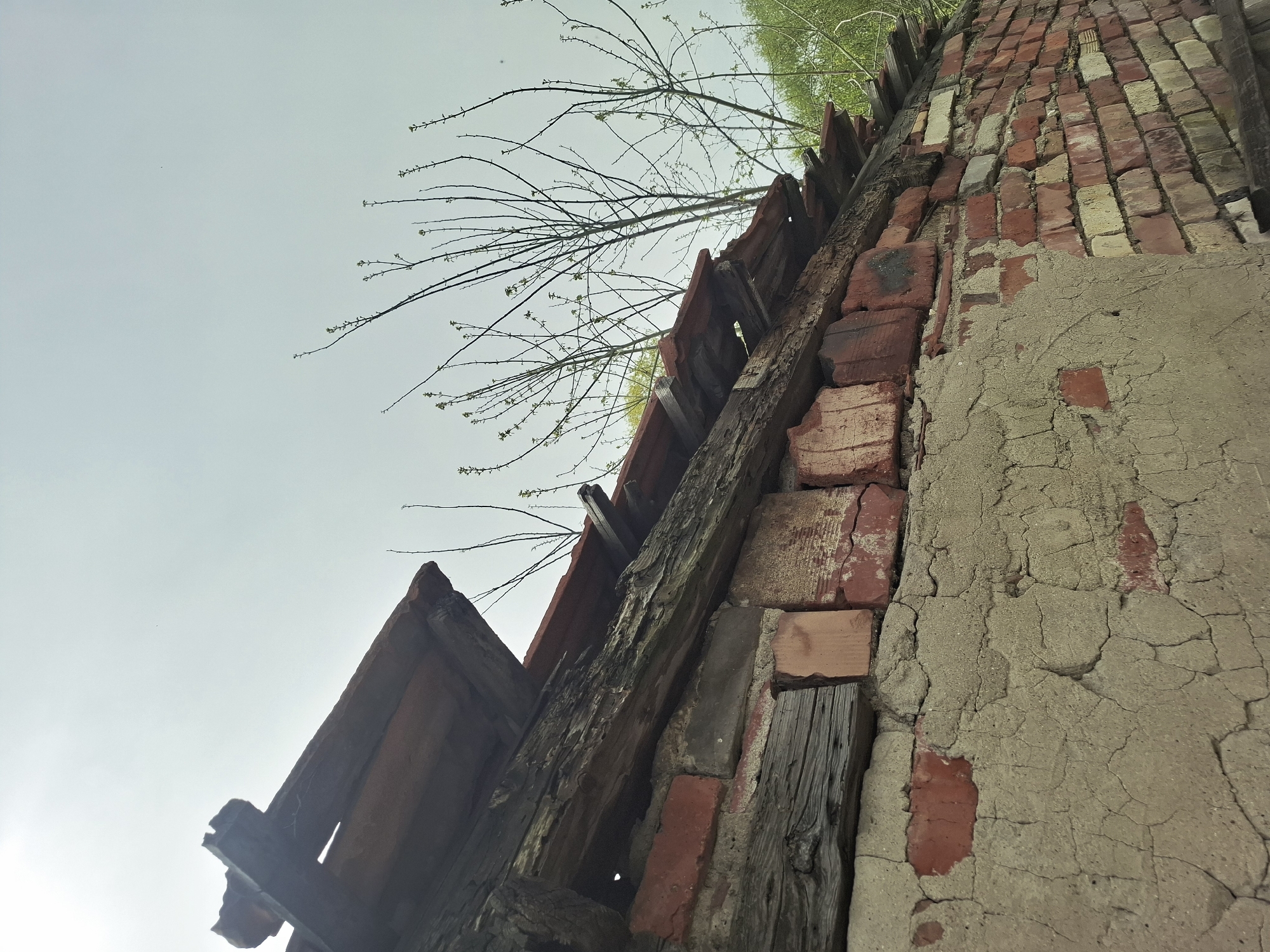 The image shows a low-angle view of a dilapidated structure with a weathered brick wall and decaying wooden supports against a light sky. The structure appears to be in a state of disrepair. There are bare tree branches visible above the structure, suggesting a natural environment encroaching on the man-made structure. There are no people visible in the image. The location is indicated as Unknown, Unknown.