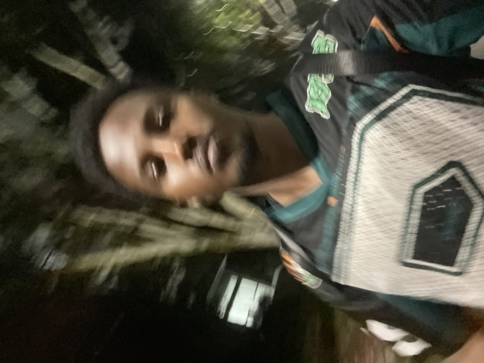 The image is a blurry selfie of a person with dark hair and skin, wearing a sports jersey with a logo. The background is dark and indistinct, possibly showing trees and a building. The image quality makes it impossible to confirm the location.