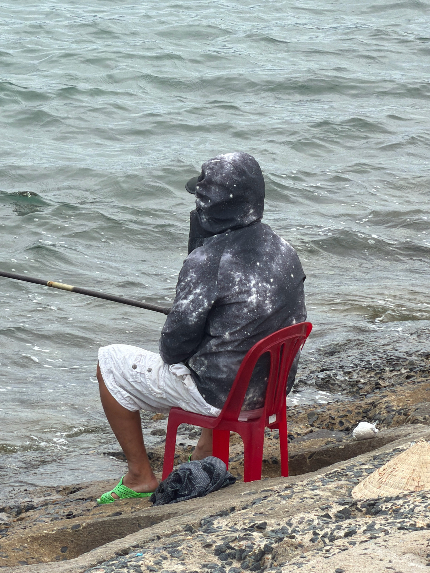 A person sits on a red plastic chair facing the ocean, holding a fishing rod. They are wearing a dark hooded jacket with white splotches and white shorts. The person has bright green flip-flops on their feet. The fishing rod extends to the left of the frame. The scene appears to be on the rocky shore of the ocean, with waves lapping at the rocks. In the background, the ocean is grey and choppy. To the right of the person, a conical straw hat rests on the rocks. A white plastic bag is also visible on the rocks near the hat. This scene is likely located along the coastline of Nha Trang, Vietnam, a city known for its beaches and fishing industry.