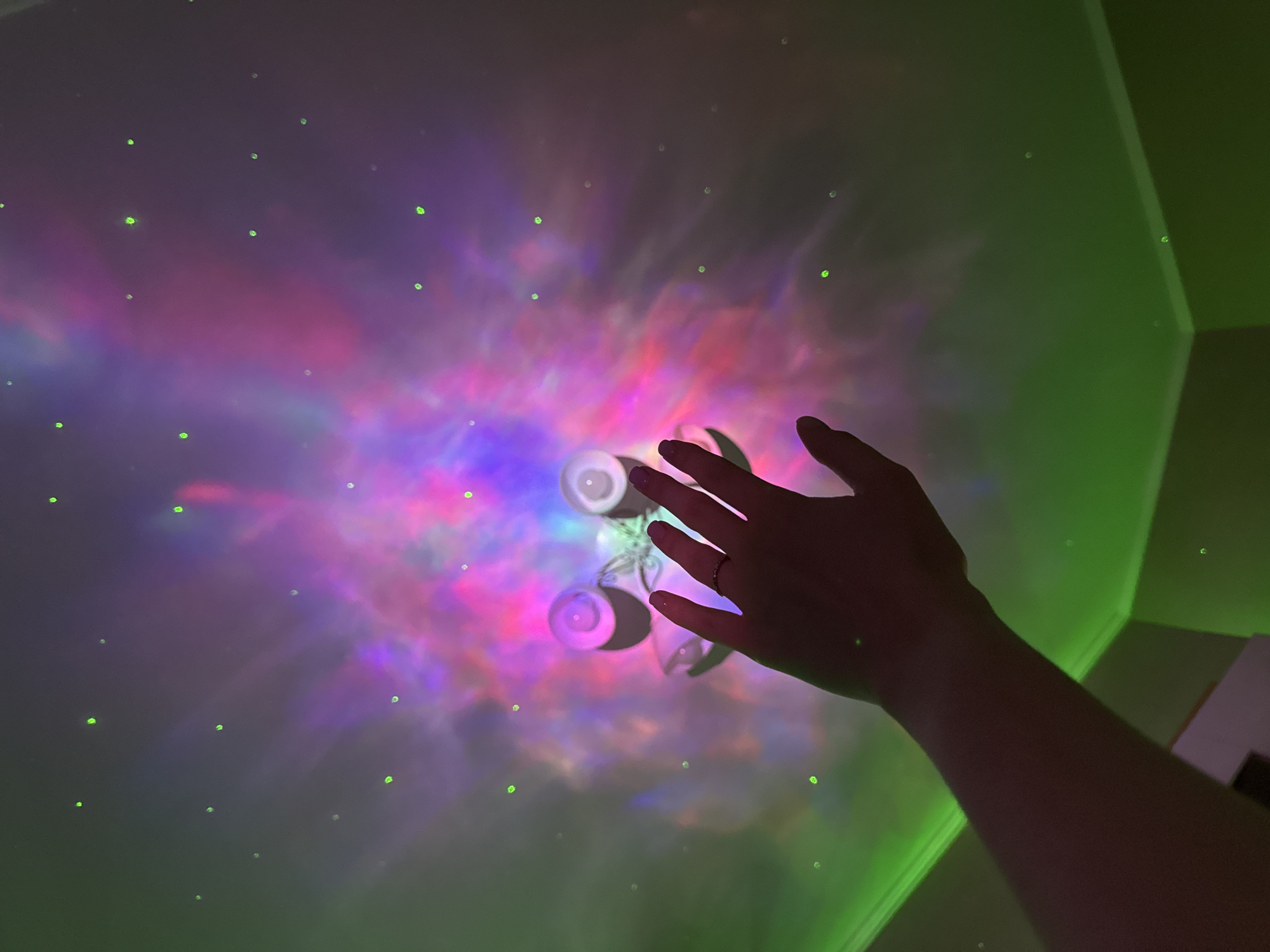 In a dimly lit room, a hand is raised towards a ceiling adorned with a vibrant projection of colorful nebulae in hues of pink, purple, and blue, interspersed with small, green, star-like lights. The hand, with fingernails painted a deep shade of purple, wears a thin ring on its ring finger. The projection emanates from a lamp shaped like a frog, its two bulbous eyes visible. A green light also casts a glow along the ceiling and down the walls, suggesting the presence of an LED strip. The environment appears to be residential, with a corner of the ceiling visible where the green light accentuates the architectural detail.