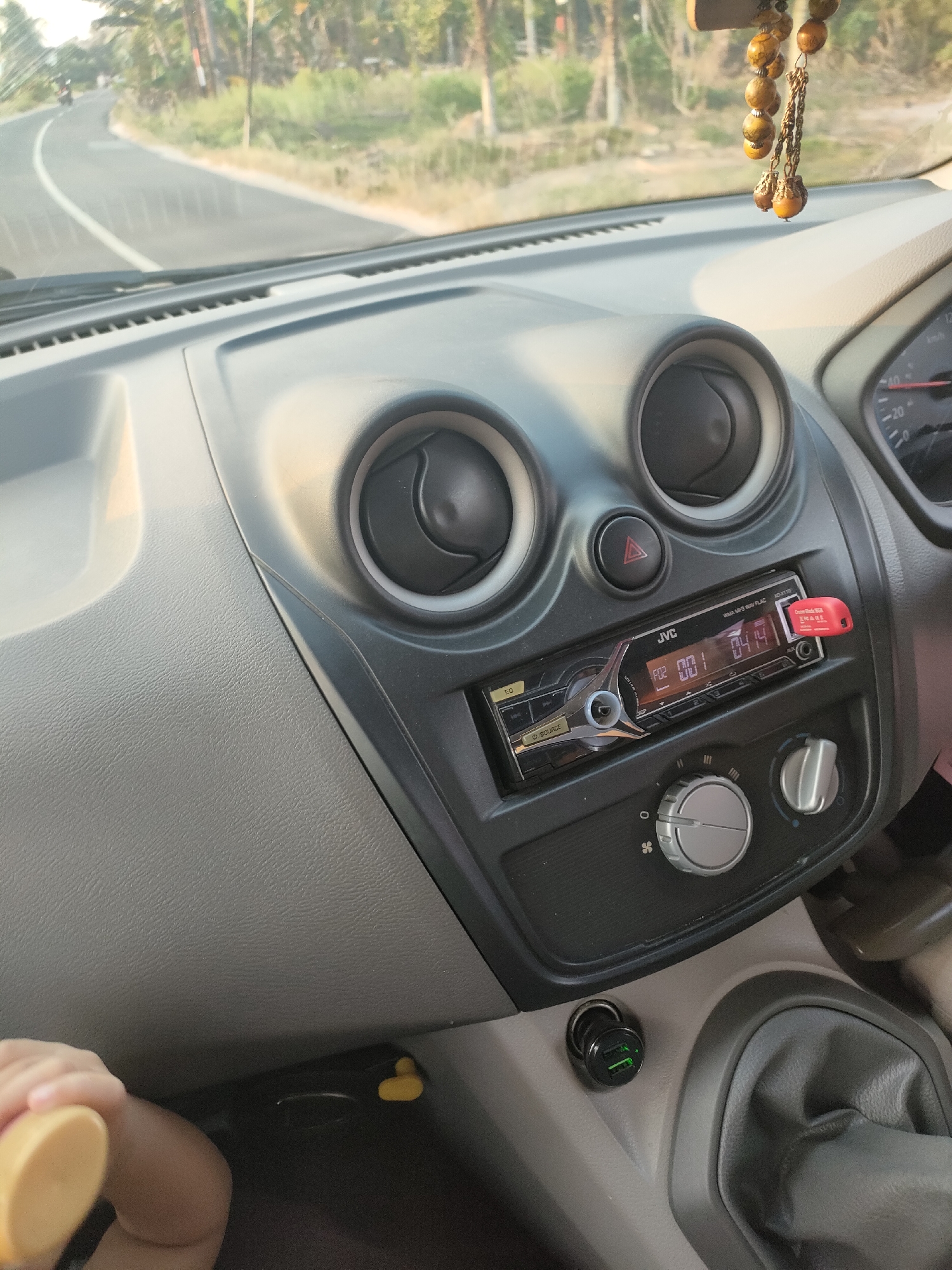 The image shows the interior of a car dashboard. A JVC car stereo is visible, with a USB drive plugged into it. There are two circular air vents above the stereo. A hazard light button is located between the vents. A car charger with two USB ports is plugged into the socket below the stereo. A person's hand is visible in the lower left corner, holding a yellow object. The car is on a road, which appears to be a rural area, with trees and vegetation visible outside the car. The road is winding and seems to be in motion. The location is stated to be Kuday, Indonesia.