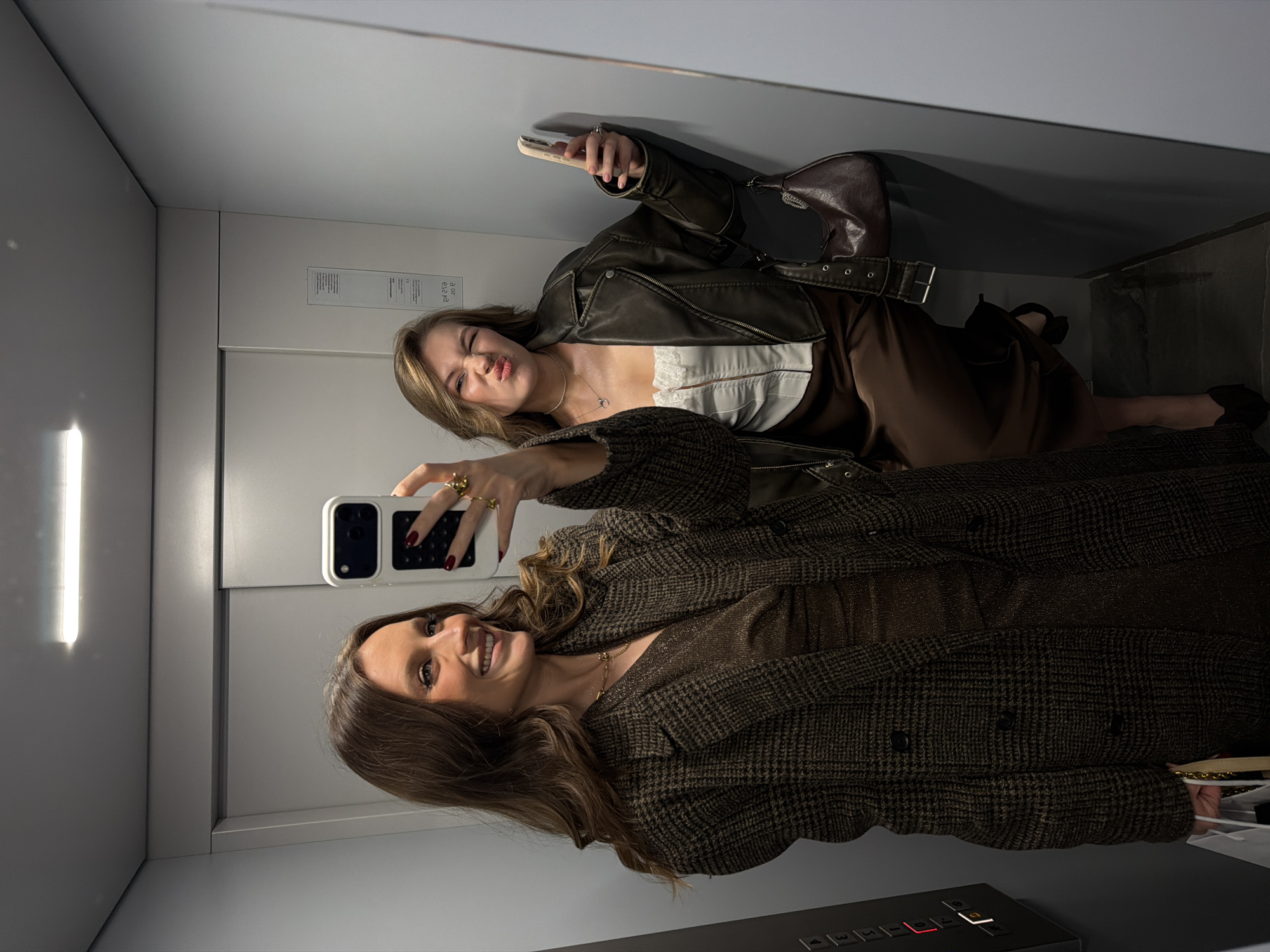 Two women are taking a selfie in an elevator. The woman in the foreground is wearing a brown plaid coat and smiling. The woman in the background is wearing a brown leather jacket and making a silly face. The elevator has a control panel with buttons. The location is an elevator in Vozdovac Urban Municipality, Serbia.