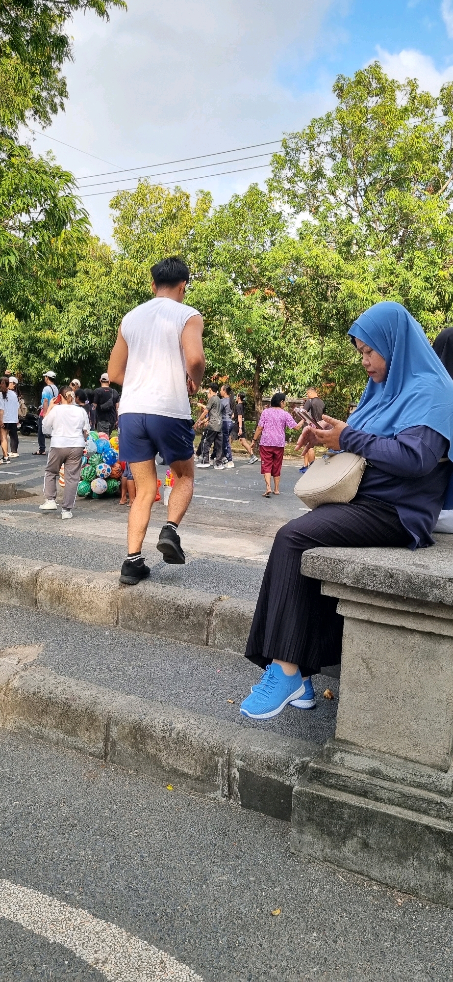 A street scene in Denpasar, Indonesia, features a group of people in the background, some appearing to be participating in an event like a fun run. In the foreground, a man in a white sleeveless shirt and dark blue shorts is running on a textured asphalt road. He is wearing black sneakers with white socks. To the right, a woman wearing a bright blue hijab and a dark blue top is seated on a concrete structure, looking at her phone. She is wearing a black pleated skirt and blue athletic shoes. In the mid-ground, a collection of colorful inflatable balloons, possibly for sale or as part of an event, are visible near the running man. The environment includes a sidewalk with stone borders, trees with green foliage, and a cloudy sky.