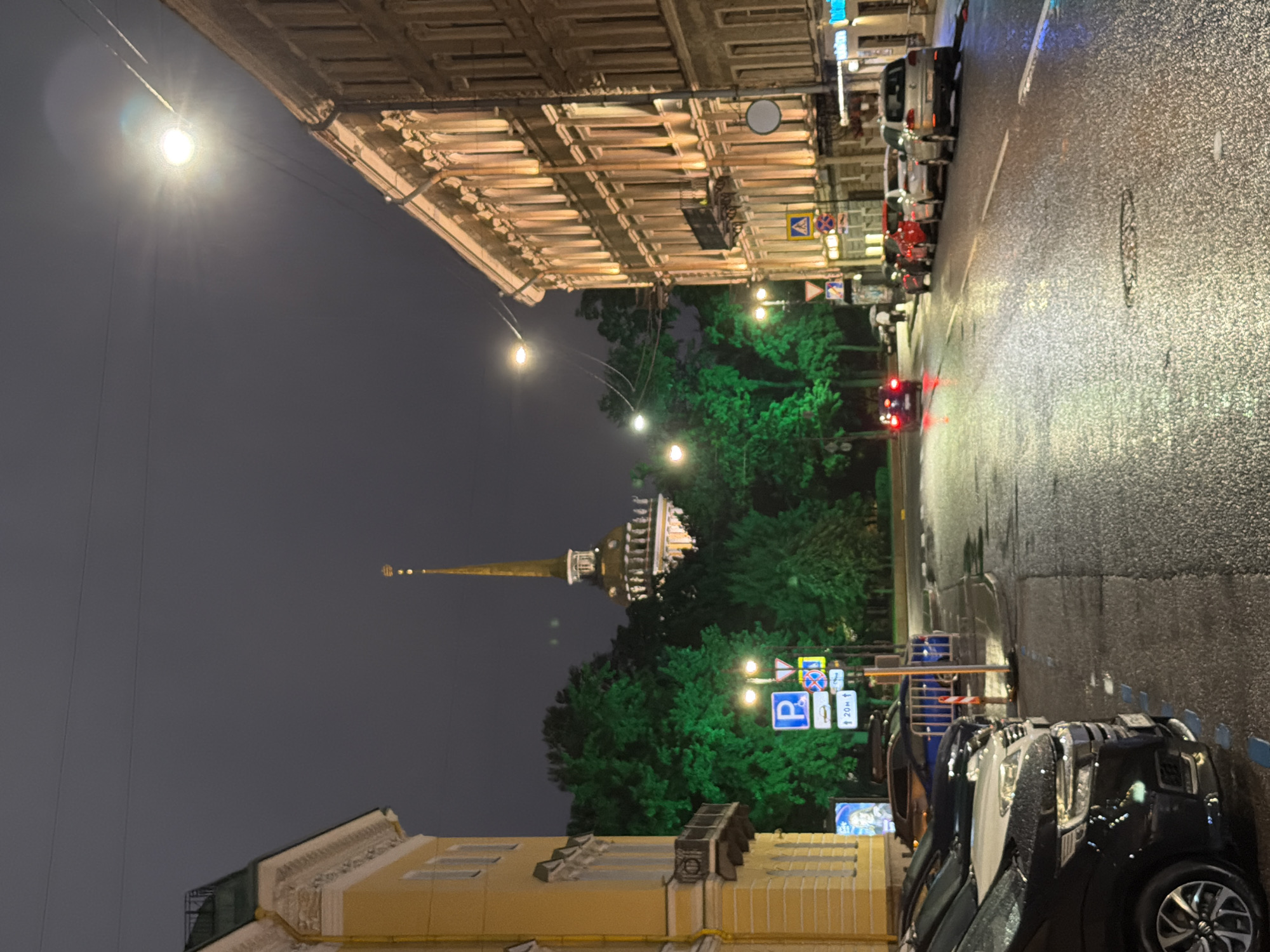 This is a nighttime, eye-level, vertical, medium-shot photograph taken from a street in Saint Petersburg, Russia, looking down a wet, asphalt road toward a tall, spired building with a golden top and illuminated windows. The sky is dark and overcast.
On the left side of the street, a tall, yellow building with decorative white trim and several windows is visible. A dark metal balcony juts out from its facade. In front of this building, a row of parked cars, including a black SUV, a white SUV, and a dark blue sedan, are parked along the curb. A blue and white parking sign is affixed to a pole. Adjacent to the cars, several trees are illuminated with bright green light, creating a stark contrast with the dark surroundings.
On the right side of the street, a multi-story building with a light-colored facade is illuminated by warm, yellow lights, highlighting its many windows and decorative architectural details. A lamppost with multiple lights is situated near the intersection. Several cars, including a silver sedan, a red car, and a white car, are parked along the right side of the road. Further down the street, a few cars are driving, their taillights visible.
The asphalt road is wet and reflective, indicating recent rain. Streetlights cast a glow on the scene, and the overall atmosphere is dimly lit. A few people are visible on the sidewalk in the distance. The spired building in the background is the Admiralty Building, a prominent landmark in Saint Petersburg.