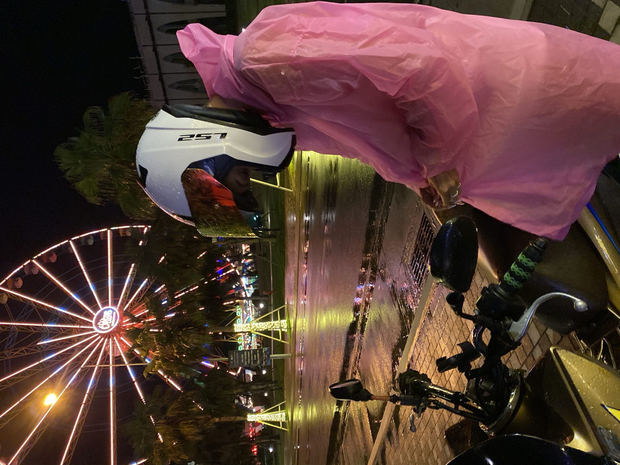 A night scene in Tien Thanh Ward, Vietnam, depicts a person wearing a pink raincoat and a white "LS2 257" helmet, possibly on or near a motorcycle. The person is bending forward, and their left hand with rings is visible holding the edge of the raincoat. The front portion of a motorcycle, including its handlebars, mirror, and a silver lever, is visible in the foreground on the right, suggesting the person might be seated on or standing next to it. The ground appears wet and reflective, indicating recent rain or ongoing precipitation.
In the background, a brightly lit Ferris wheel with red and white lights stands prominently against the dark night sky. Several palm trees are also visible, interspersed with other illuminated structures and small buildings, suggesting a fairground or amusement park setting. The overall atmosphere is dark, with artificial lights casting reflections on the wet surfaces.