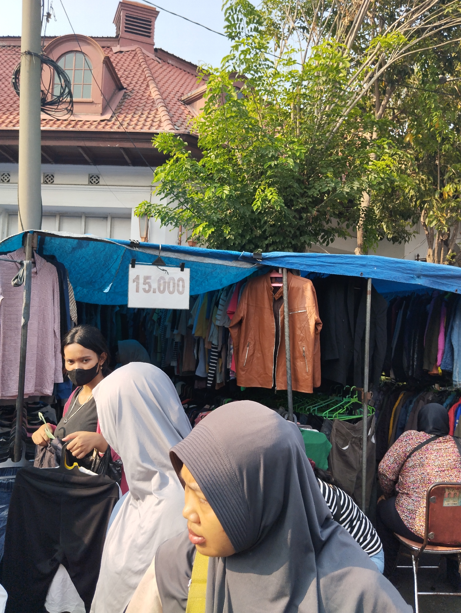 A street market in Surabaya, Indonesia is depicted. A clothing stall displays various garments under a blue tarp. A sign with "15.000" is visible. In the foreground, three people are present. One person, wearing a black face mask, is looking at the camera. Another person is wearing a light grey hijab, and a third person is wearing a dark grey hijab and facing away from the camera. A woman with a floral patterned shirt is seated on a chair in the background. The surroundings show a street with trees and a building with a red tiled roof, suggesting an urban setting.