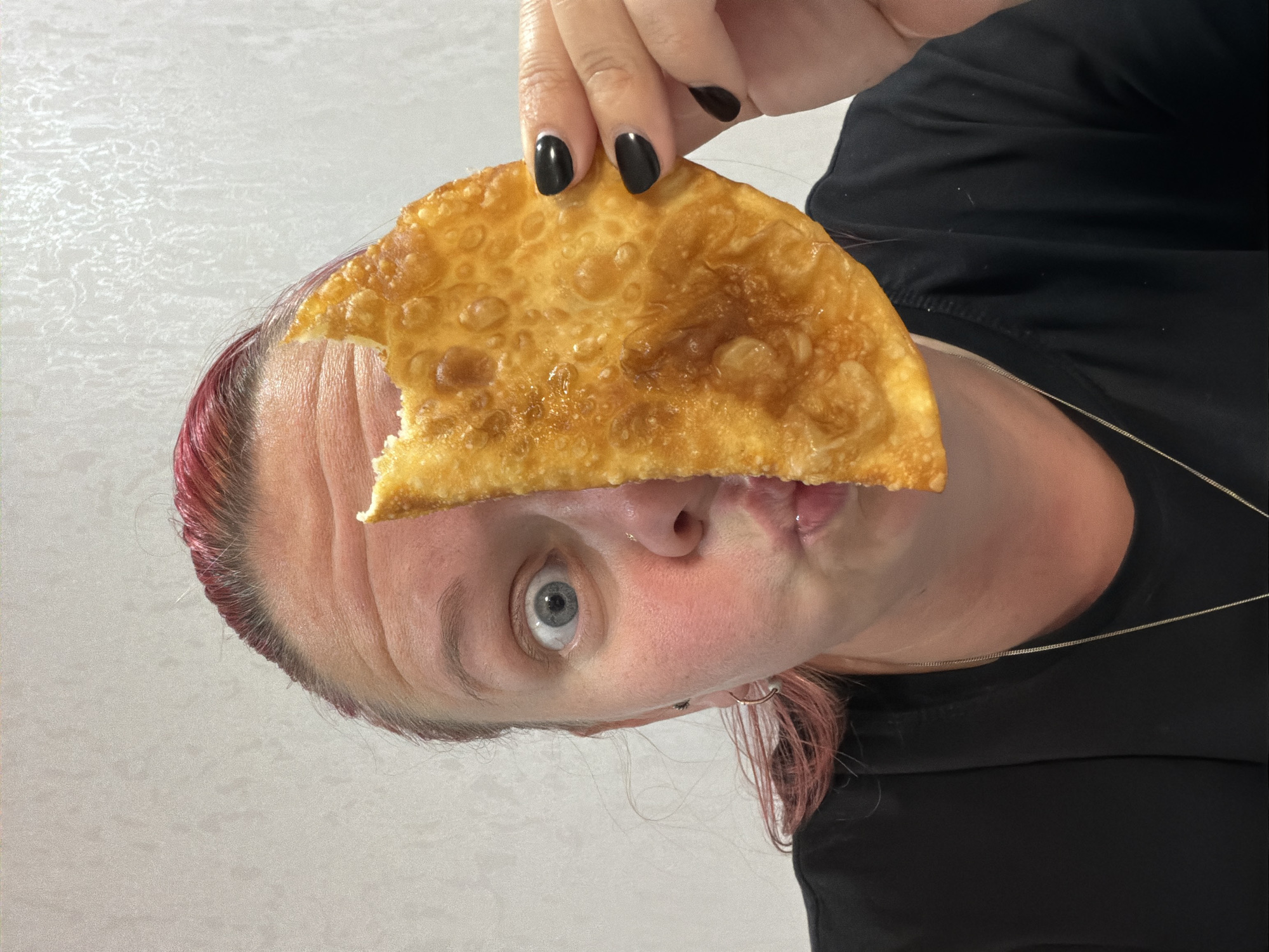 An over-the-shoulder, medium shot captures a person's face from the nose up, with their right eye and mouth visible. The person's hair is dyed a bright pink. They are holding a golden-brown, fried pastry with a bite taken out of it in their right hand, obscuring the left side of their face and forehead. The pastry is round and has a bumpy, textured surface. The person's left eye is wide open, and their lips are pursed. They are wearing a black t-shirt and a silver necklace. The background is a plain, textured off-white wall.