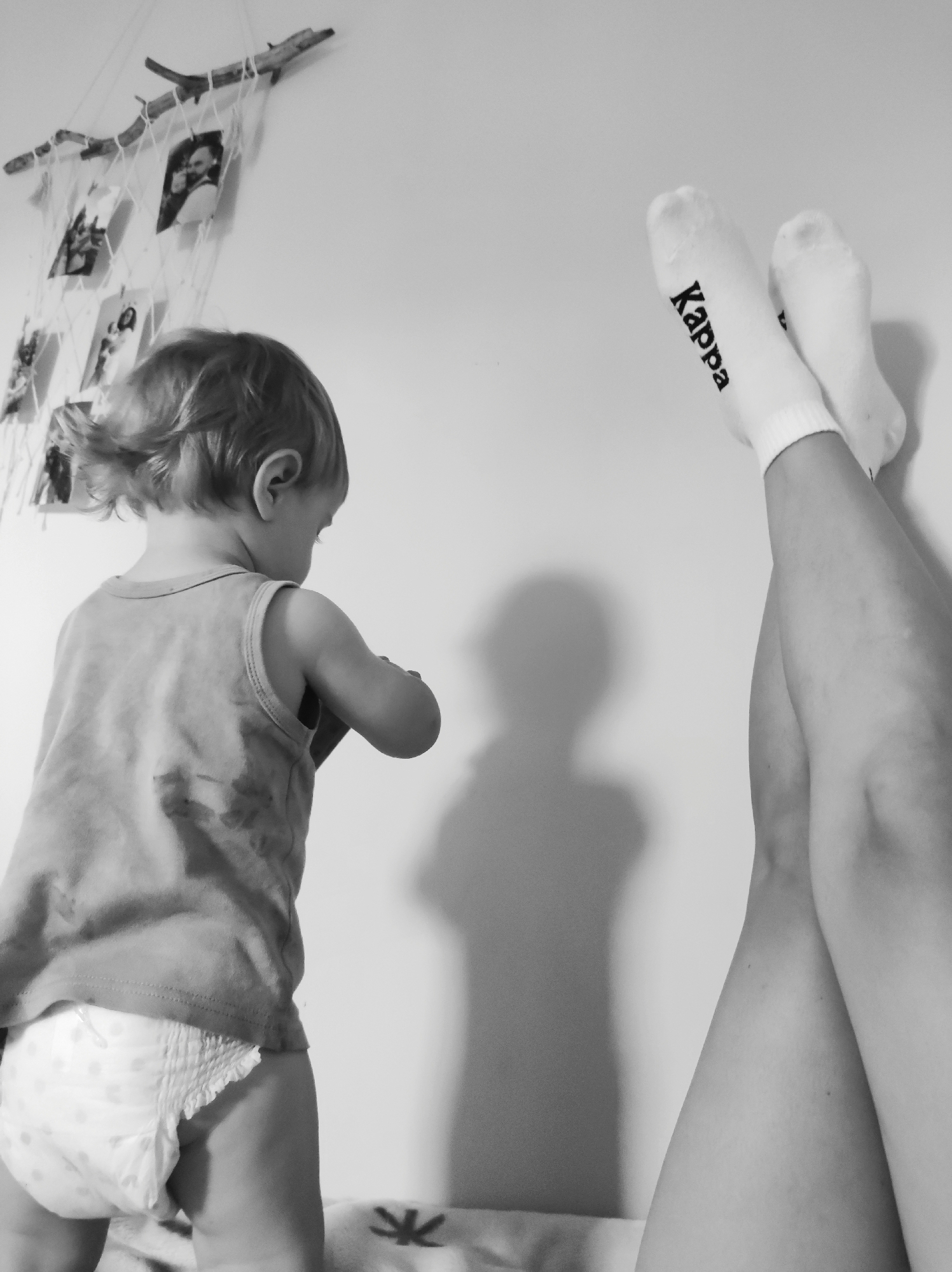 A high-angle, black and white photograph captures a moment between a baby and an adult indoors. On the left side of the frame, a young child, seen from the back, stands facing a wall. The child has short, light-colored hair and is wearing a sleeveless top and a diaper with visible patterns.
To the right, an adult's legs are raised against the wall, with the feet, covered in white socks bearing the "Kappa" logo in black lettering, visible at the top right of the image. A shadow of a person, likely the adult whose legs are visible, is cast on the wall in the center of the frame.
In the upper left corner, a decorative item hangs on the wall. It appears to be made of two pieces of wood connected by string or netting, from which several small photographs or postcards are suspended. The lighting suggests an indoor setting, possibly daytime given the shadow, but no specific time of day can be definitively determined. The overall atmosphere is quiet and domestic.