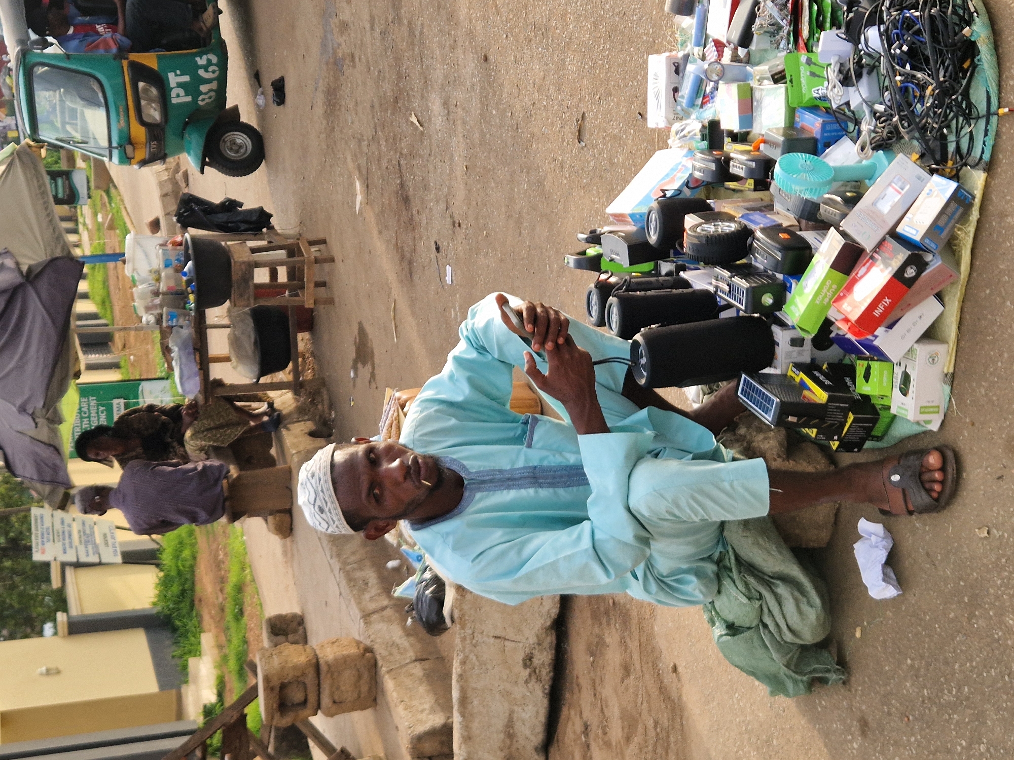 The image depicts a street scene in Jos, Nigeria, near a market or roadside vendor area. In the foreground, a man wearing a light blue traditional outfit and a white cap is seated on a green fabric pile next to a collection of electronics. He is holding a small electronic device in his hands. The electronics include speakers, portable fans, power banks, and various cables. In the background, there are more stalls with goods, a tricycle, and two other individuals sitting on a bench. A table with more items, possibly food or drinks, is also visible. The ground is paved with asphalt and concrete. The overall setting appears to be an outdoor marketplace.