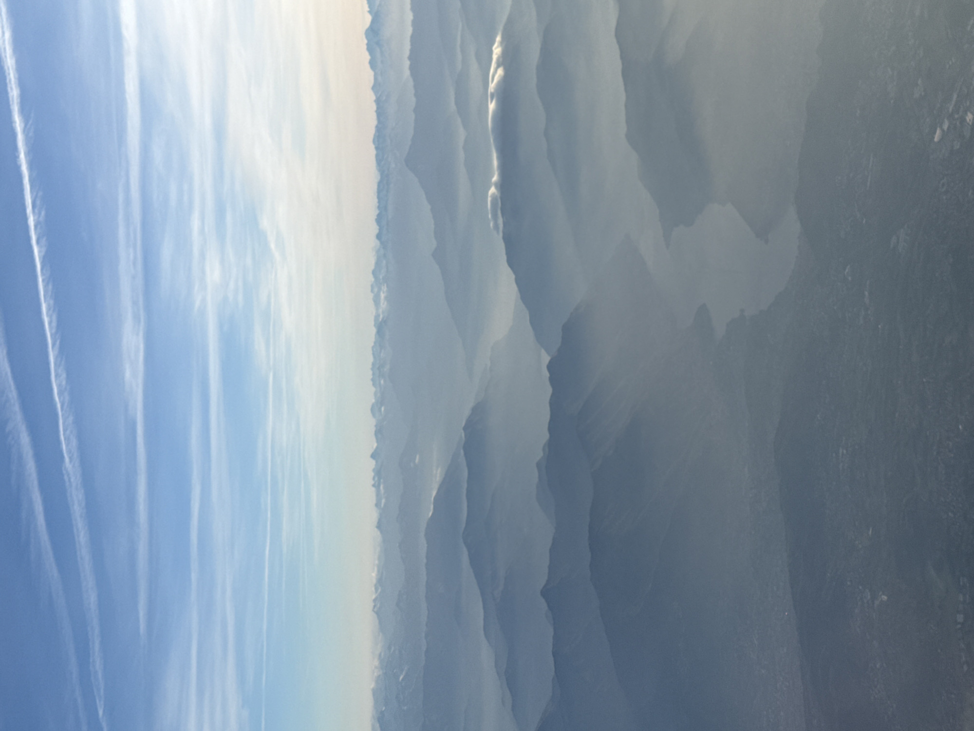 The image depicts a landscape with a blue sky partially obscured by thin clouds on the left side. Contrails are visible in the sky. The right side of the image reveals a layered terrain of mountains or hills fading into a muted gray, possibly due to atmospheric haze or distance. The photograph appears to have been taken from a high altitude, potentially from an aircraft. The location is not identifiable in the image.