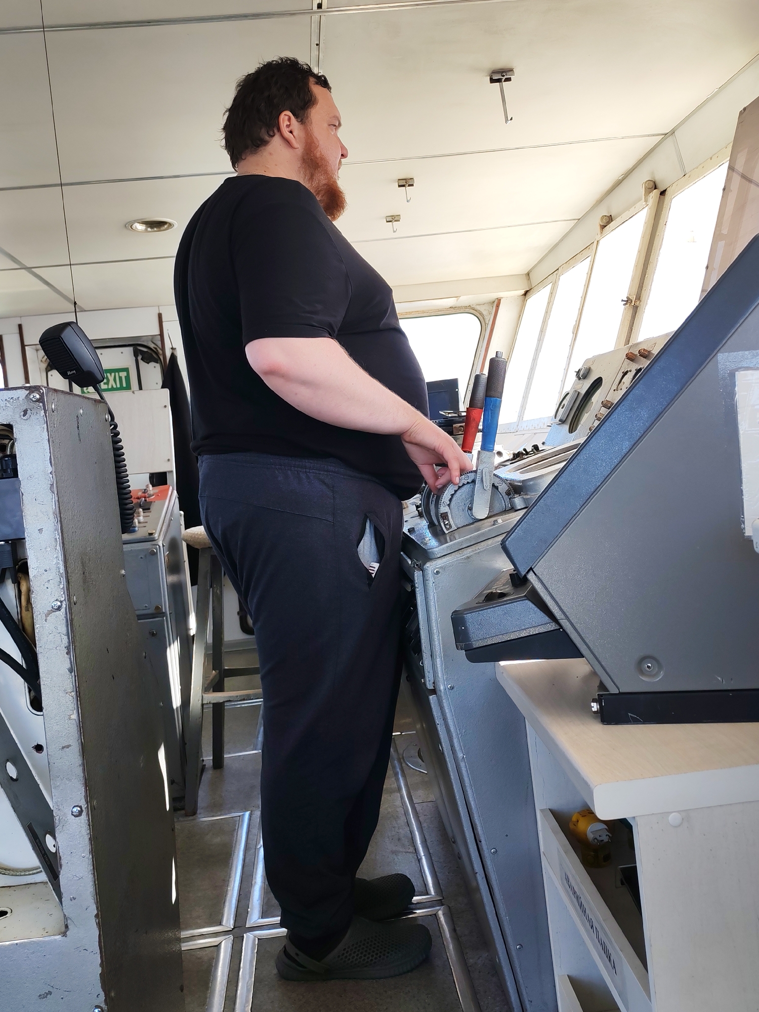 The image shows a man standing in what appears to be the bridge of a ship. He is fair-skinned with a red beard, wearing a black t-shirt and dark-colored sweatpants, and dark shoes. He is standing facing to the right of the frame. His hand is on a mechanical lever of some sort, part of the ship's controls. The ship's instruments are visible, mostly gray, with a white surface underneath. Light is coming in through the windows. There is an "EXIT" sign visible in the background.