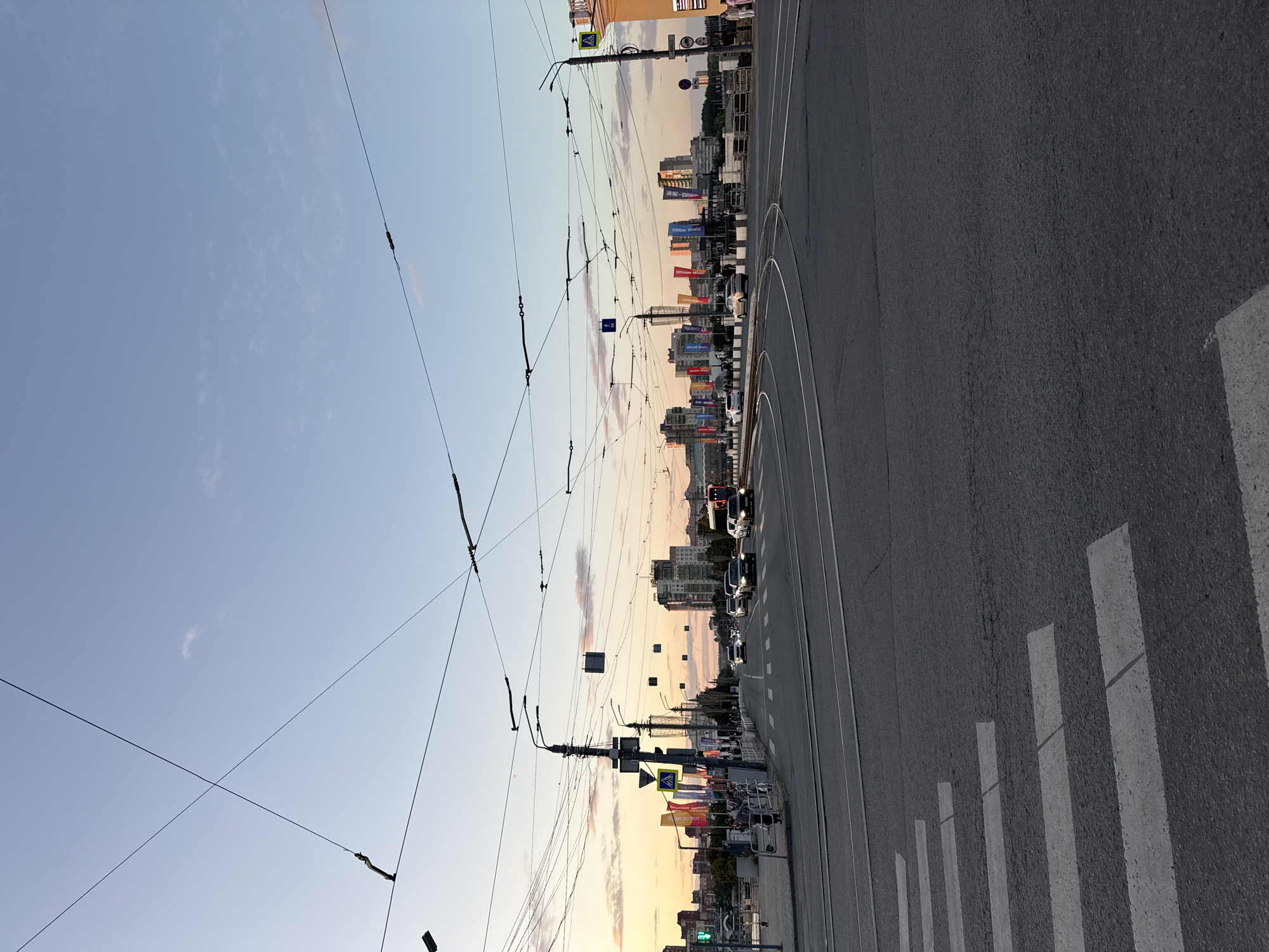 The image depicts a wide, paved intersection in an urban setting, likely a city like Chelyabinsk, Russia, given the architectural style and presence of overhead power lines for trams or trolleybuses. The sky is a soft gradient of twilight colors, transitioning from pale blue at the top to a warm, peachy hue near the horizon. In the background, a cityscape is visible, characterized by several modern, multi-story buildings. Along the horizon, a line of colorful flags, possibly promotional or celebratory banners, are displayed prominently. The foreground features a pedestrian crossing with white stripes painted on the asphalt. Several cars are driving on the road, and there are streetlights and traffic signs throughout the scene. The overall impression is of a busy urban environment at dusk.