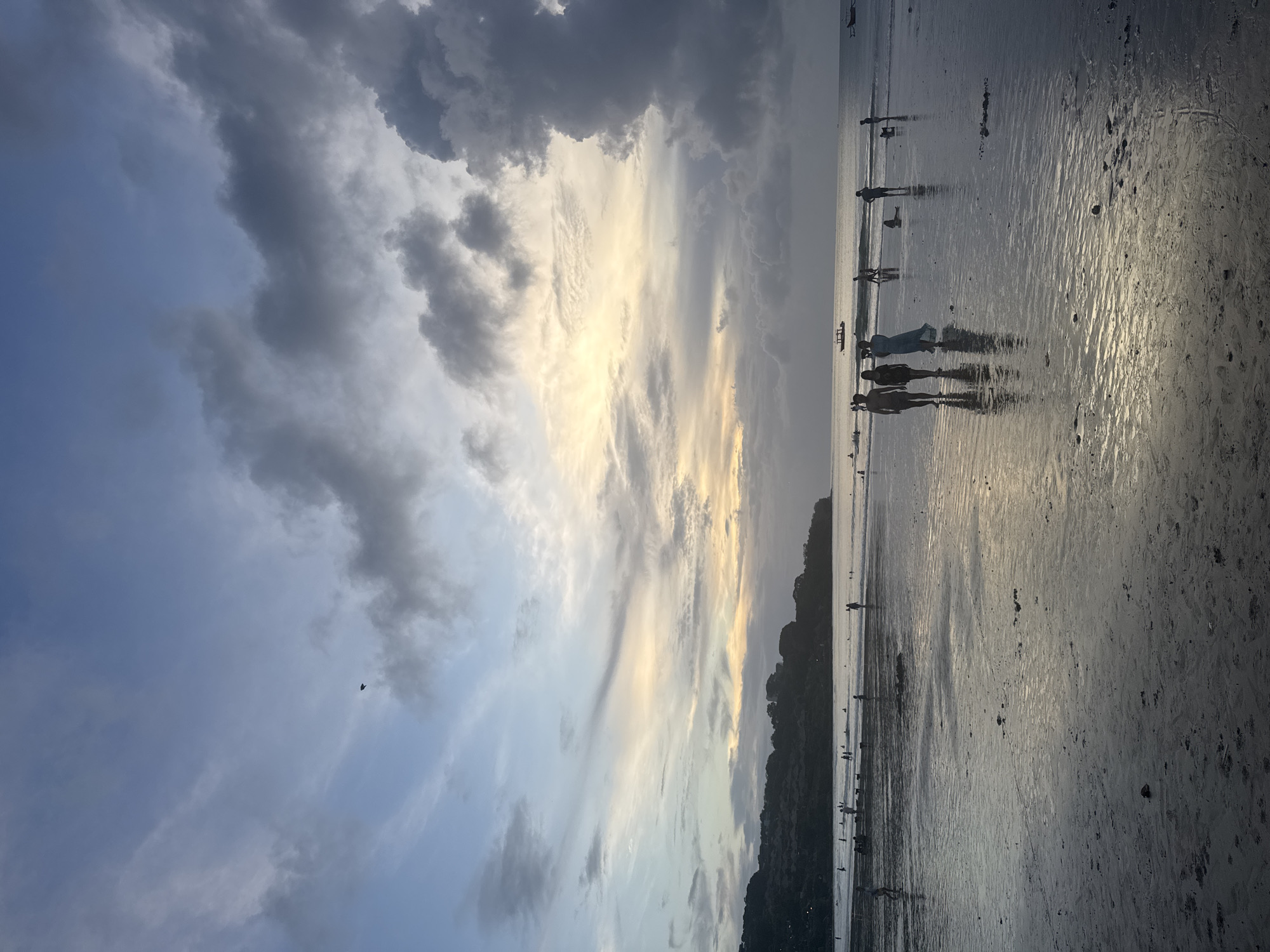 The image captures a coastal landscape during either sunrise or sunset, characterized by dramatic lighting and a serene atmosphere. The sky dominates the upper portion, featuring a striking mix of dark grey, white, and light blue clouds. A brilliant band of golden light stretches horizontally across the middle of the sky, indicating the low position of the sun. In the lower half, a wide expanse of wet sand or very shallow water reflects the sky, creating shimmering patterns and highlighting the texture of the surface.
Numerous people are visible as dark silhouettes along the water's edge, most appearing to be standing or walking in the shallow water. Their forms are indistinct due to the backlighting. Several small, elongated boats are also silhouetted further out on the water. To the right, a dark, undulating landmass, possibly an island or coastline covered in dense vegetation, extends along the horizon. A small, dark, V-shaped object, possibly a bird, is faintly visible against the lighter sky in the upper left. The overall impression is one of peaceful natural beauty at the "golden hour." There is no visible text in the image.