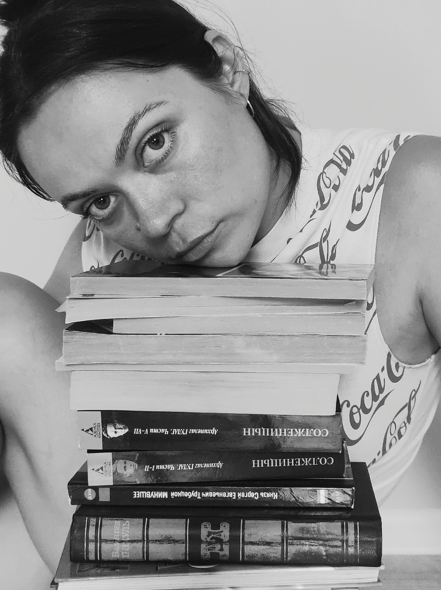 A grayscale portrait captures a woman with dark hair and a Coca-Cola patterned shirt leaning on a stack of books. Her gaze is direct, with a slightly melancholic expression. The books are stacked unevenly, with the top ones being plain and the lower ones having visible titles in Cyrillic, including works by Solzhenitsyn. The environment appears simple, with a light-colored backdrop.