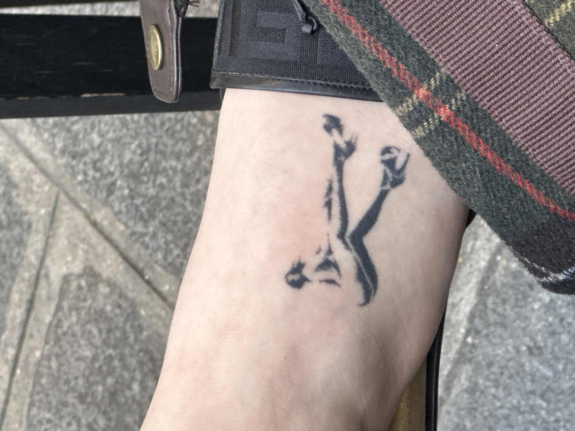 A close-up shot shows a person's lower leg, adorned with a black tattoo of what appears to be a stylized depiction of the Eiffel Tower. The leg is positioned against a background of grey stone pavement, typical of Parisian sidewalks. Adjacent to the leg, there are elements of clothing: a section of a green and red plaid fabric, and part of a black textured material with a brown leather detail and a gold button. The lighting is natural and diffused, suggesting an outdoor setting during the daytime.