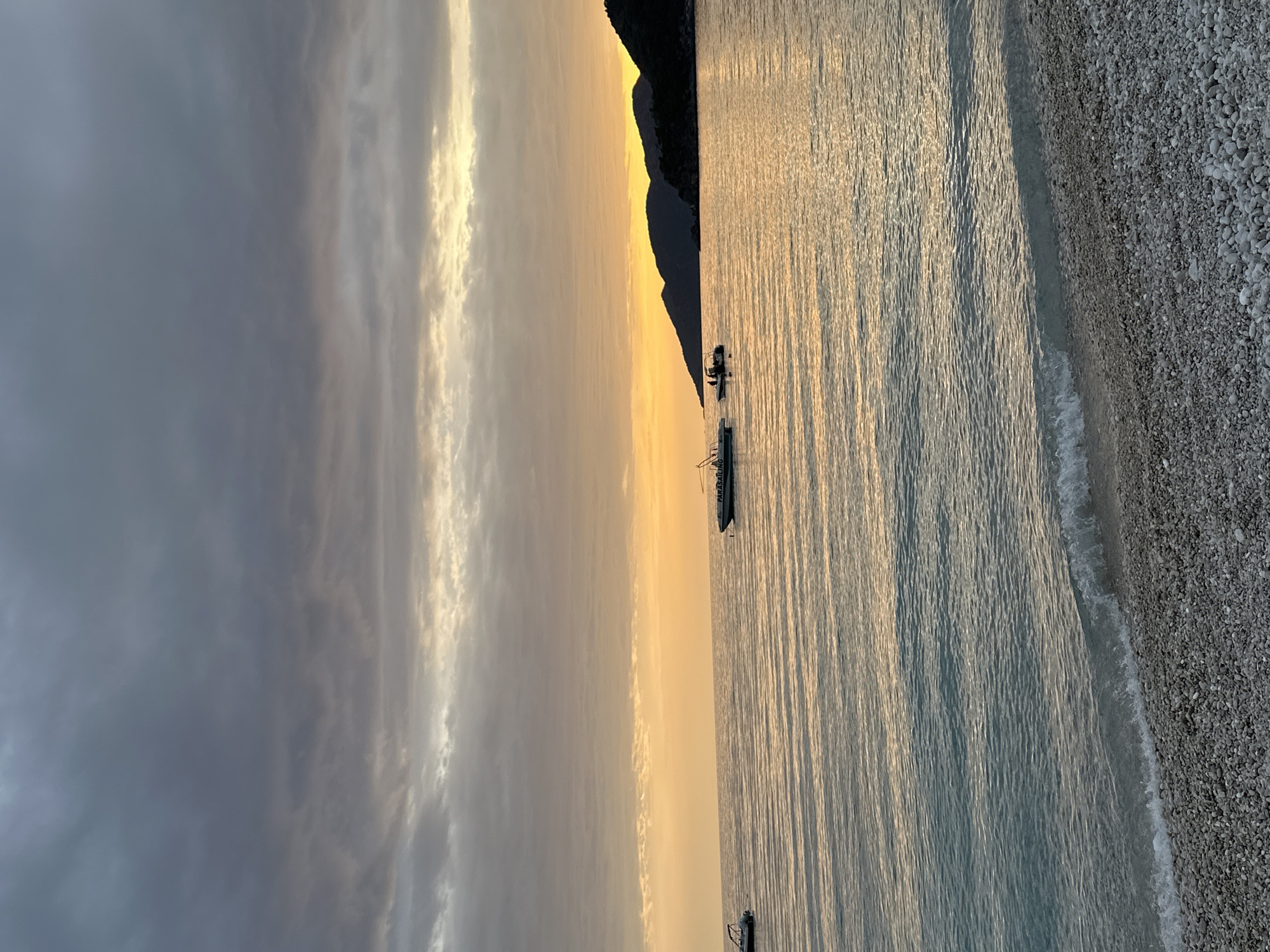 The image shows a shoreline scene in Fethiye, Turkey at sunset. The water is calm with a glassy surface reflecting the yellow and orange colors of the setting sun. Small waves lap against a pebble beach on the right side of the frame. In the distance, a dark mountain silhouette stands against the bright sunset sky. Two boats are visible in the water near the mountain. The left side of the frame is dominated by gray, cloudy sky.