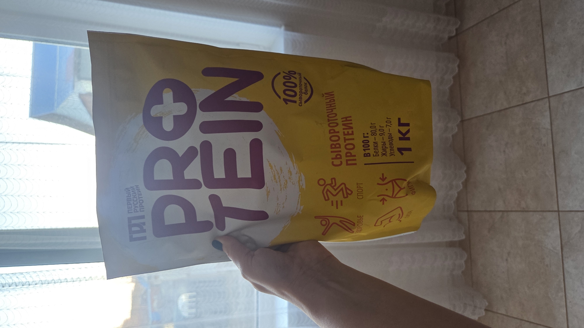 A person is holding a yellow bag of "ProTein" whey protein. The bag indicates "First Russian Protein" and a weight of 1kg. Nutritional information includes 80g of protein, 9g of fat, and 7g of carbohydrates per 100g. The bag also has icons representing sports, health, strength, and figure. A window is visible in the background, suggesting an indoor location. The image appears to be taken in Chaikovsky, Russia, as the product description is in Russian.