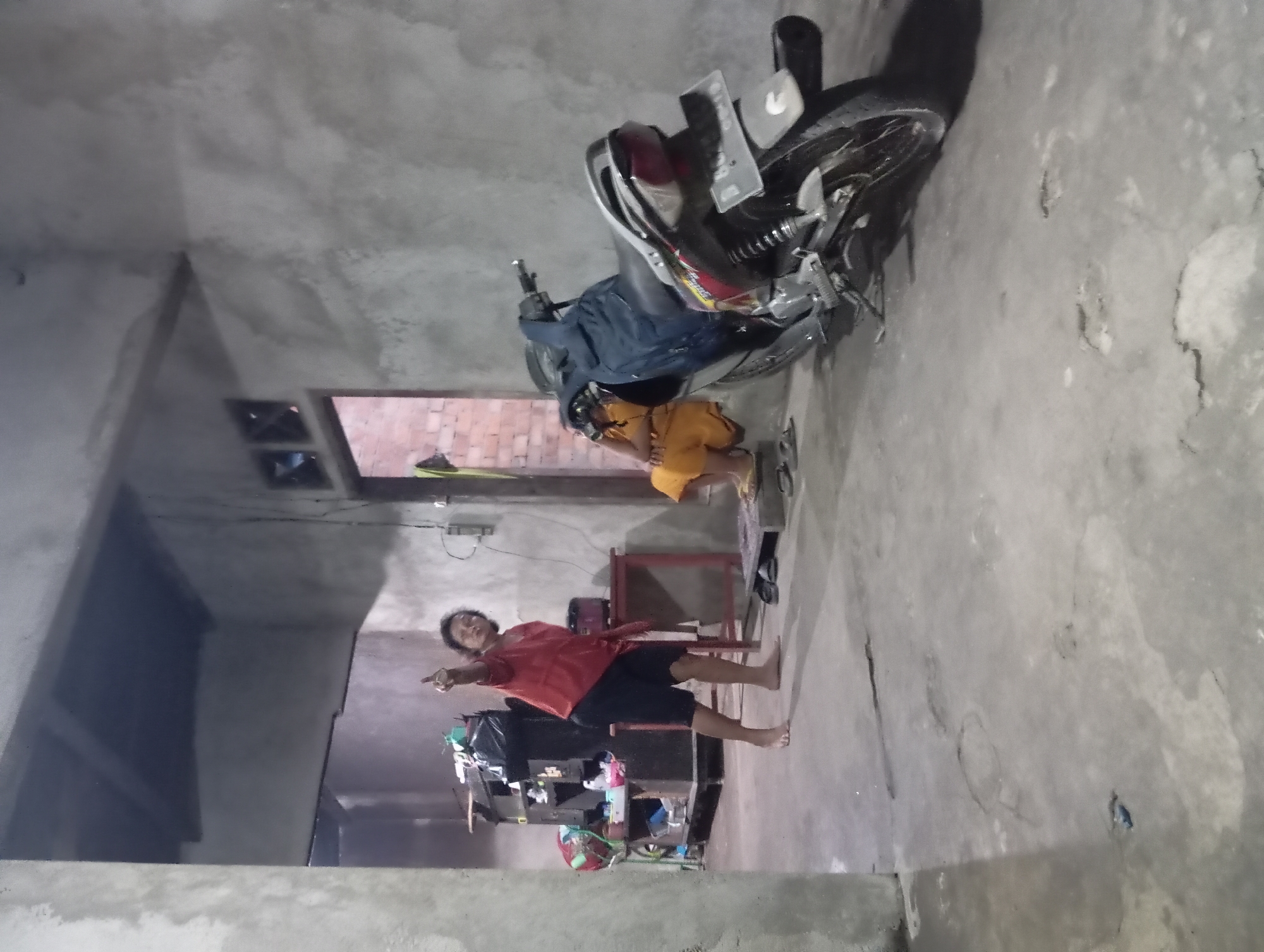 The image depicts an interior scene in what appears to be a modest dwelling in Rejosari, Indonesia. The environment is sparsely furnished with unfinished concrete walls and a concrete floor. In the foreground, a dark-colored motorcycle is parked. To the left, a person wearing a red shirt and black shorts stands barefoot, pointing upwards with their right index finger. This individual's pose suggests they are directing attention or making a gesture. To the right and slightly behind the motorcycle, another person is seated on the floor, wearing an orange garment. The background reveals a doorway leading to another area, a piece of furniture with various items stored on it, including a large black trash bag, and a small red table. The lighting is dim, creating shadows and an overall subdued atmosphere. The scene is one of everyday life within a simple home.