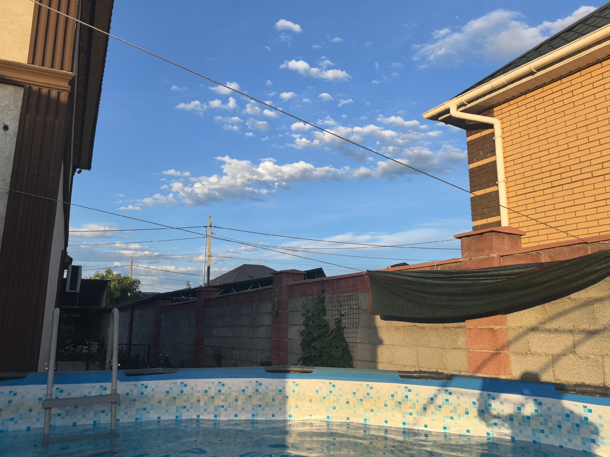 The image depicts an outdoor swimming pool area on a sunny day. In the foreground, the edge of the pool is visible, lined with a mosaic of blue and white tiles. A silver ladder extends into the pool, suggesting it's ready for use. The background features a sturdy fence, a brick house with a gutter system, and part of another building. The sky is a vibrant blue, dotted with fluffy white clouds, indicating pleasant weather. The lighting suggests it is late afternoon or early evening, with the sun casting long shadows. The scene evokes a sense of leisure and relaxation, likely in a residential setting. There is no visible text in the image. The location is Taldykorgan, Kazakhstan.