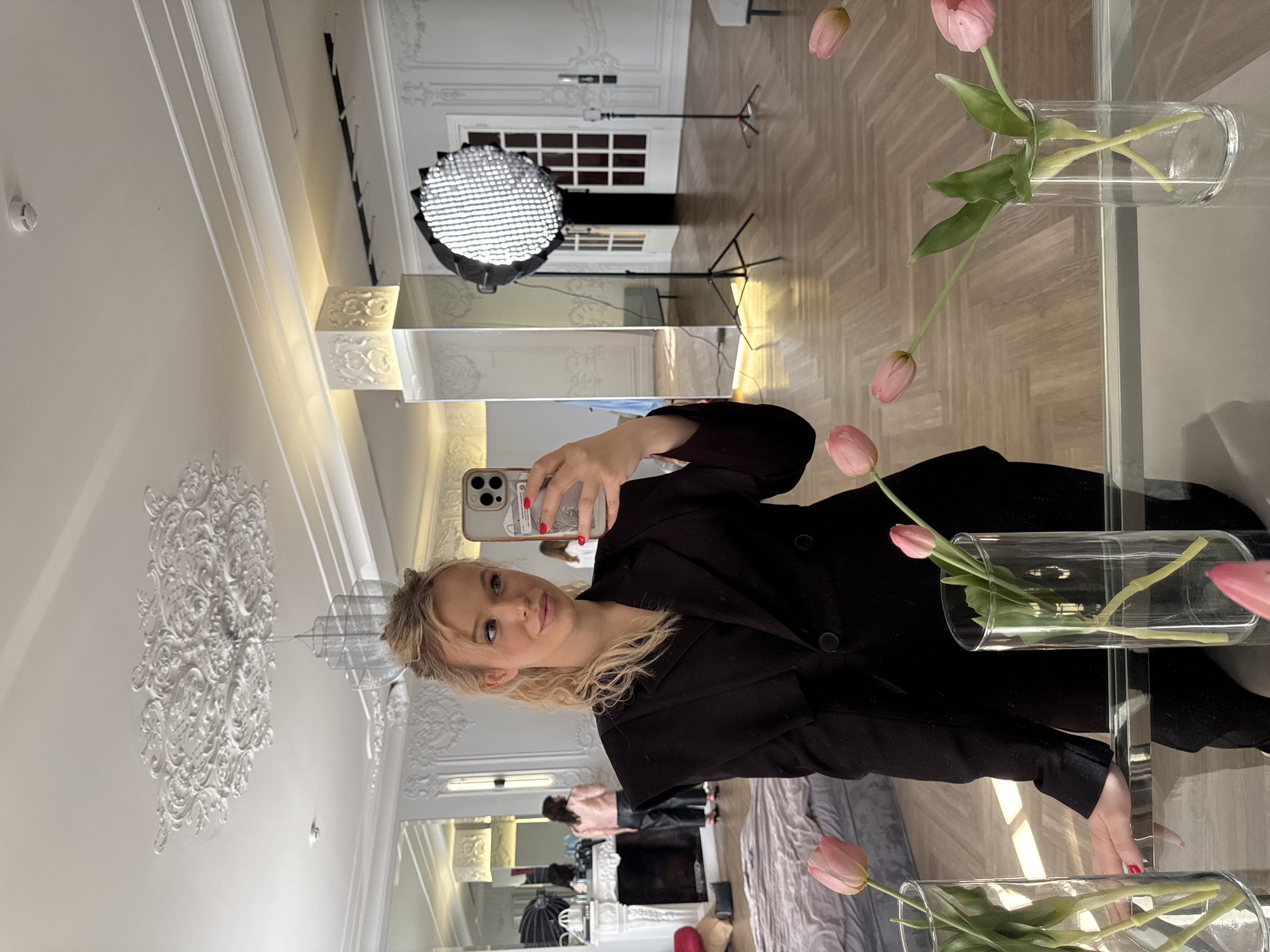 A young woman with blonde hair pulled up, wearing a black blazer and pants, takes a selfie in a large mirror. She is in a well-lit, spacious room with ornate white moldings on the walls and ceiling, reminiscent of classical St. Petersburg architecture. In the reflection of the mirror, two other women are visible; one in a pink jacket and black pants standing near a fireplace, and another in the background. A large photographic softbox light is positioned on a stand, and several glass vases with pink tulips are placed on a reflective surface in the foreground. The room has a parquet floor and furniture, including a bed, that suggest it might be a studio or a well-appointed apartment. The overall impression is one of a professional photoshoot or a stylish interior.