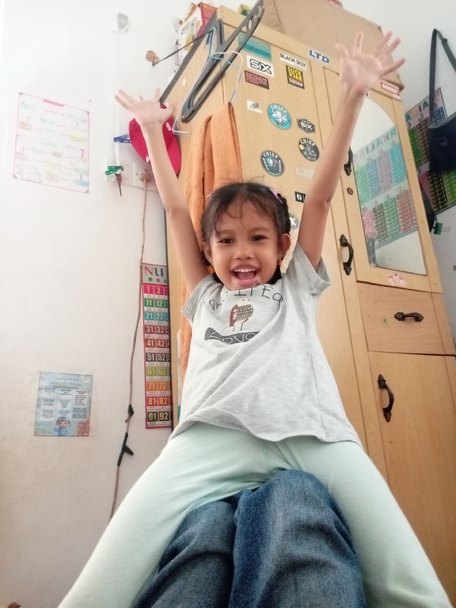 The image depicts a young girl seated on someone's lap, with her arms raised in the air. She is wearing a light grey t-shirt with a graphic print and light blue pants. Her facial expression is joyful, with an open mouth and wide eyes. Behind her, a wooden cabinet with several drawers and a mirror is visible. Various stickers and posters are affixed to the cabinet and the wall, including a large chart with numbers and what appears to be a school schedule. On the wall to the left, another poster is visible, along with electrical outlets and wires. The environment suggests an indoor setting, possibly a bedroom or a common living area in a residential dwelling. The overall impression is one of playful activity and familial interaction. The presence of Indonesian text on some of the posters and stickers suggests the location is in Indonesia. Given the context of the question, and the visual cues, this scene is likely situated in a home in Batam, Indonesia.