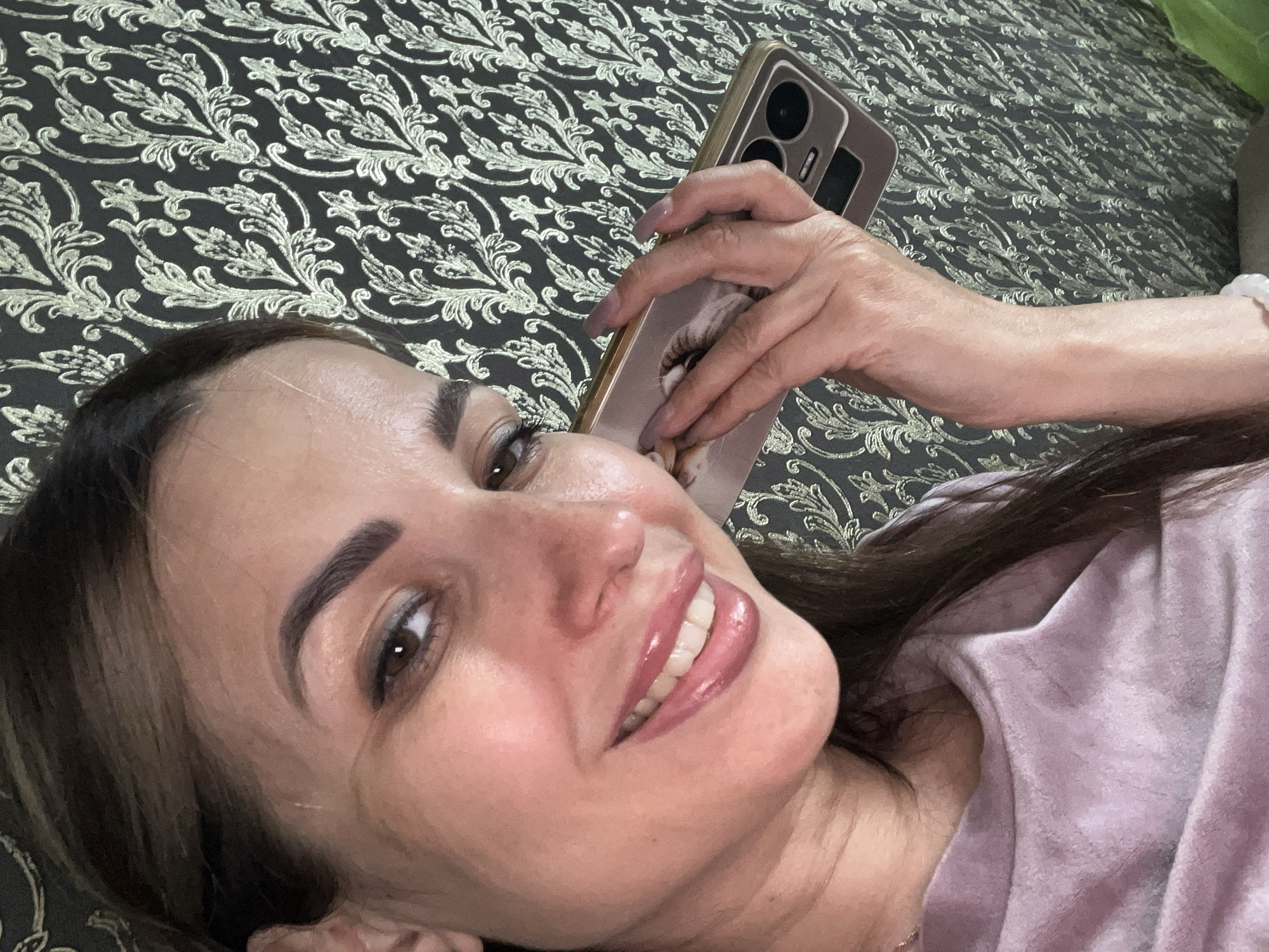 A woman with long, dark brown hair is taking a selfie while holding a smartphone to her ear. She is smiling and wearing a light purple shirt. The background is a dark wallpaper with a golden floral pattern. Her left hand is holding the phone, and her right arm is visible.