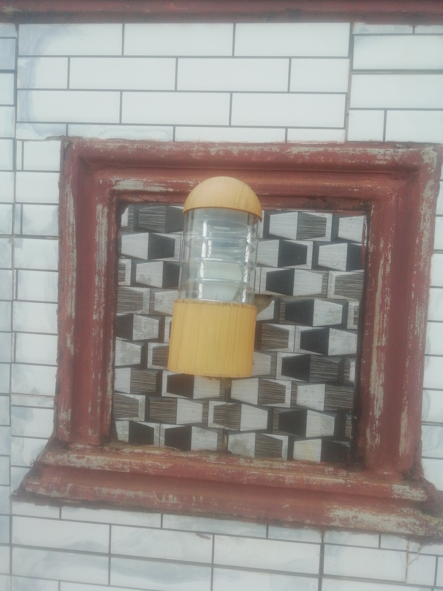 The image depicts an outdoor light fixture mounted within a recessed, red-painted frame on a white brick wall. The light fixture has a cylindrical clear glass body with a yellow wood-like top and bottom. The background within the frame features a decorative tile pattern consisting of alternating black and white 3D rectangular shapes. The scene is located in Ikorodu, Nigeria.