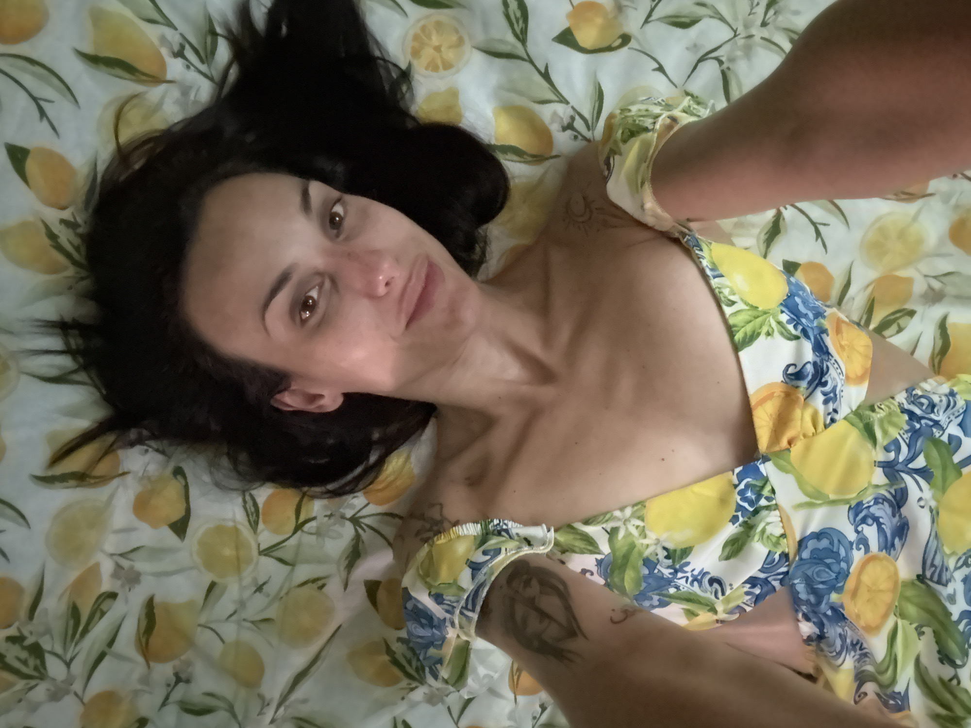 A woman lies on a lemon-printed sheet or bedspread. Her dark hair is spread around her head. She is wearing a lemon-printed dress with blue and white accents. She has tattoos on her arms and shoulder. The location is not specified in the image. There is no indication it is in Pleven, Bulgaria.
