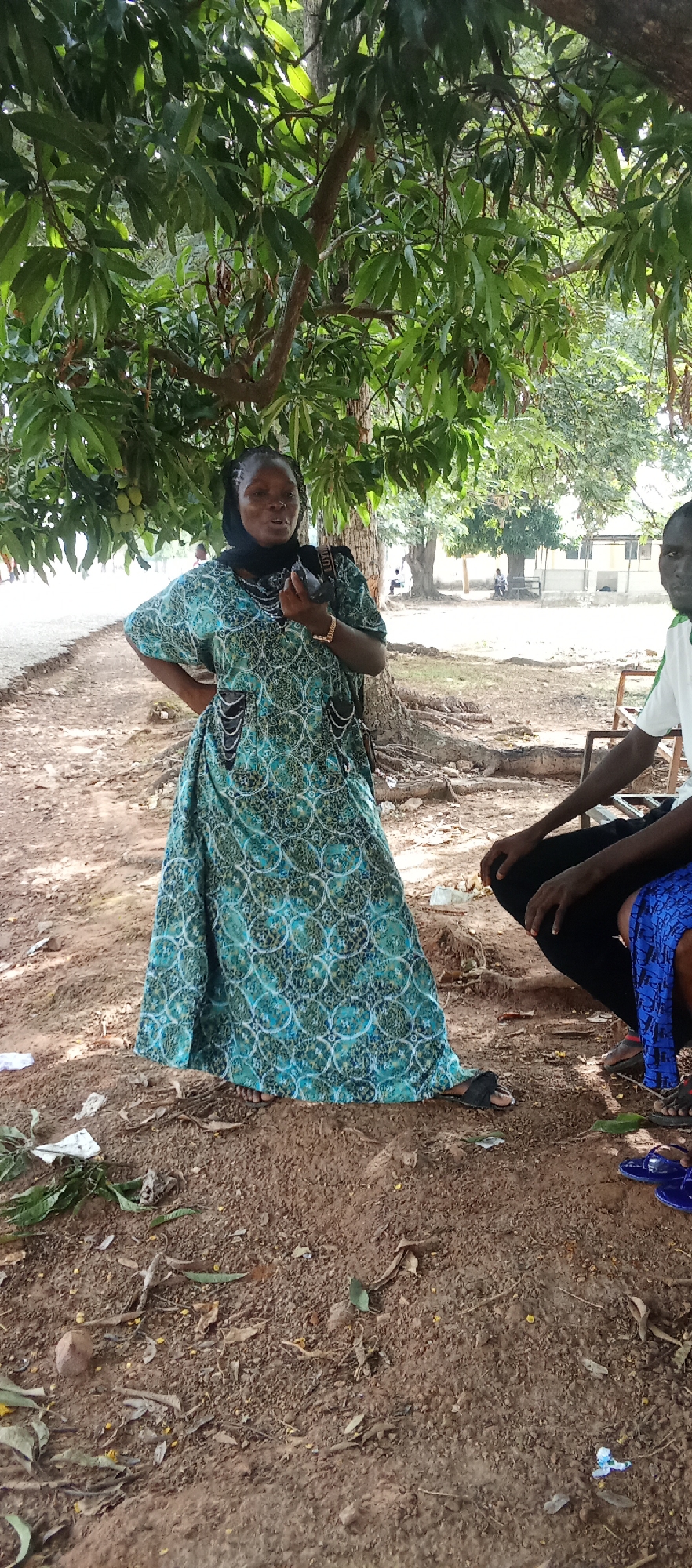 A woman in a blue patterned dress and black head covering stands beneath a tree with green leaves, likely a mango tree, given the presence of mangos. She has her hand on her hip. To her right, partially visible, is a man seated; his attire includes black pants and a shirt with a white and green stripe on the sleeve. Blue sandals are visible on the ground near him. The ground is reddish-brown dirt with scattered leaves, debris, and what appears to be litter. In the background, there is a building and figures sitting on benches. The location is Akwanga, Nigeria.
