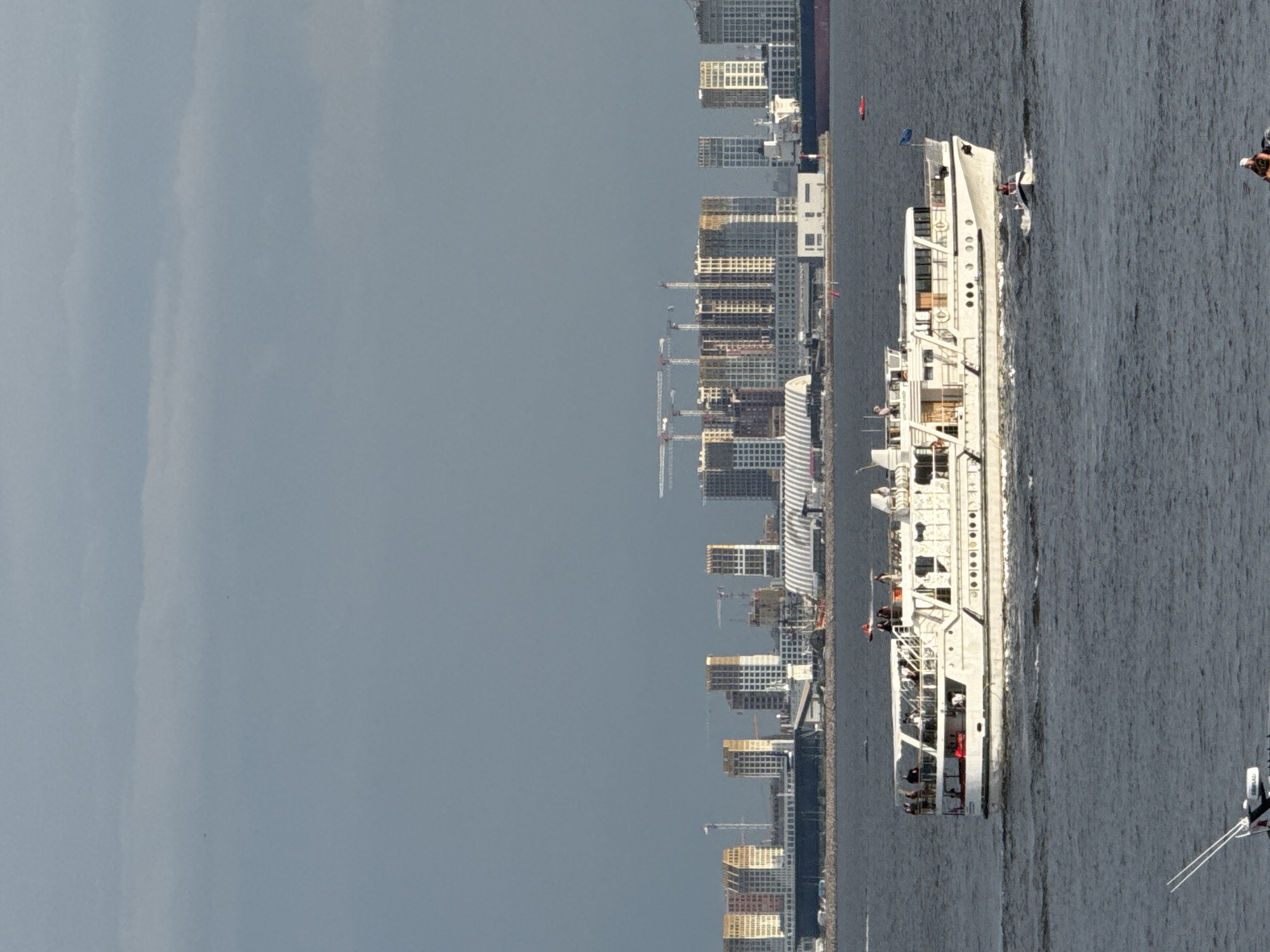 The image depicts a large, white, multi-decked boat with numerous people visible on its decks, cruising on dark, choppy water. The boat is moving from left to right in the frame, leaving a wake behind it. In the background, a dense urban skyline is visible, characterized by numerous tall, modern buildings and construction cranes, suggesting an ongoing development project. A red buoy is also present in the water near the city skyline. Further to the right, a portion of another, smaller boat with what appears to be antennae or a radar system is visible at the edge of the frame. The sky is overcast with a dull gray color, indicating possibly cloudy weather. The overall scene suggests a daytime view of a busy waterfront in a city. The orientation of the image is rotated 90 degrees counter-clockwise from what is typically considered upright.