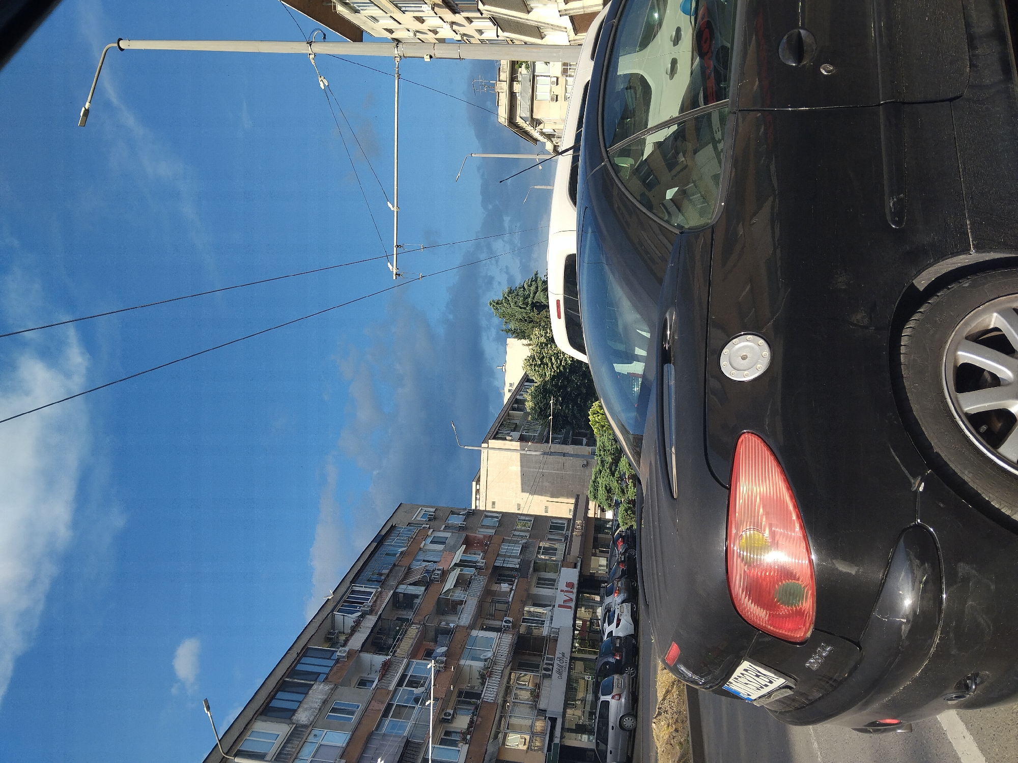 The image shows a street scene in Stara Zagora, Bulgaria. In the foreground, there is a parked black car, a Peugeot 206, angled towards the right of the image. The car is parked on the right side of the road. Its rear light and rear portion of the vehicle are visible. Adjacent to it is a white van, with part of its back visible.
Behind the cars and across the street, there is a multi-story residential building. The building has a number of windows and balconies. There is a sign on the ground floor of the building with some text, including "Ivis". Other parked vehicles are visible between the buildings and the black car, partially obscured.
Overhead, there are utility poles and wires stretching across the sky, which is blue with some clouds. The scene appears to be taken during daylight hours.